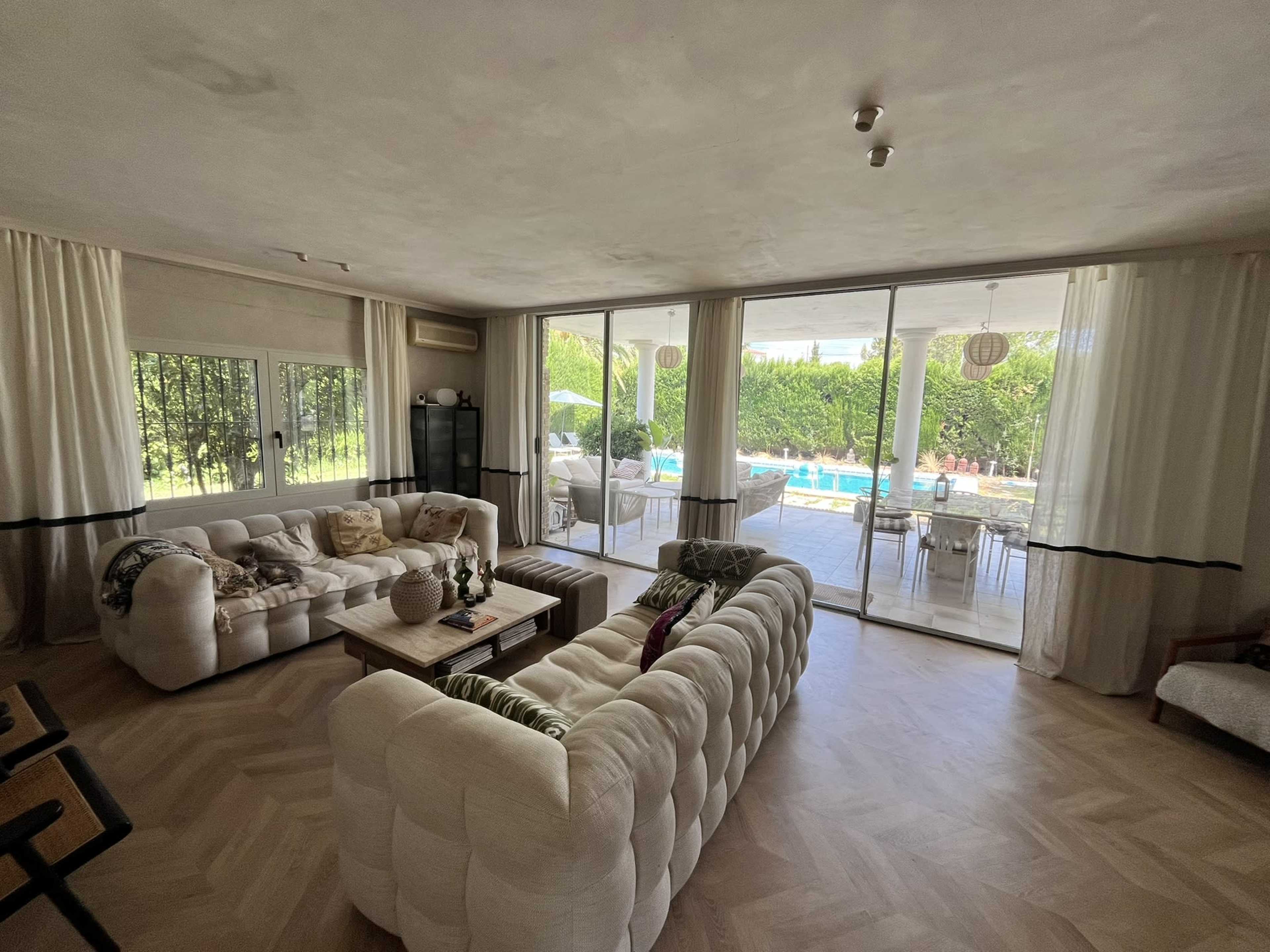 The image shows a modern living room with a large window overlooking a patio and pool area, featuring white sofas and a light wooden floor.