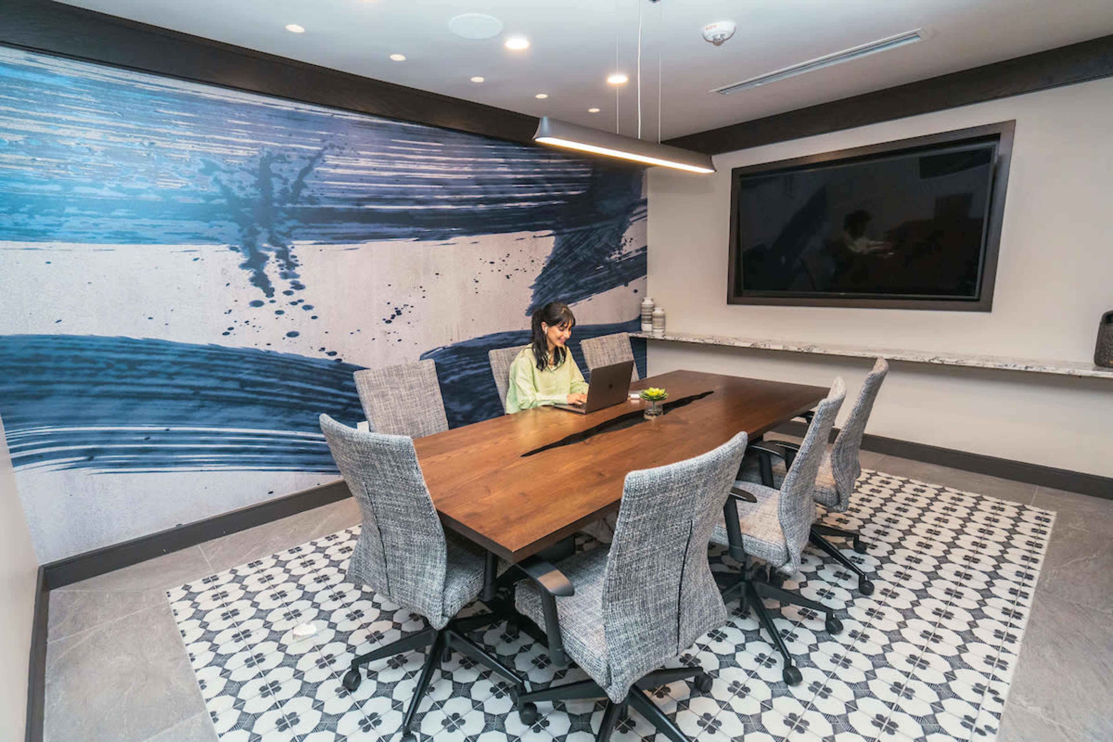 A woman is working on a laptop at a wooden table in a meeting room with a blue abstract mural on the wall.