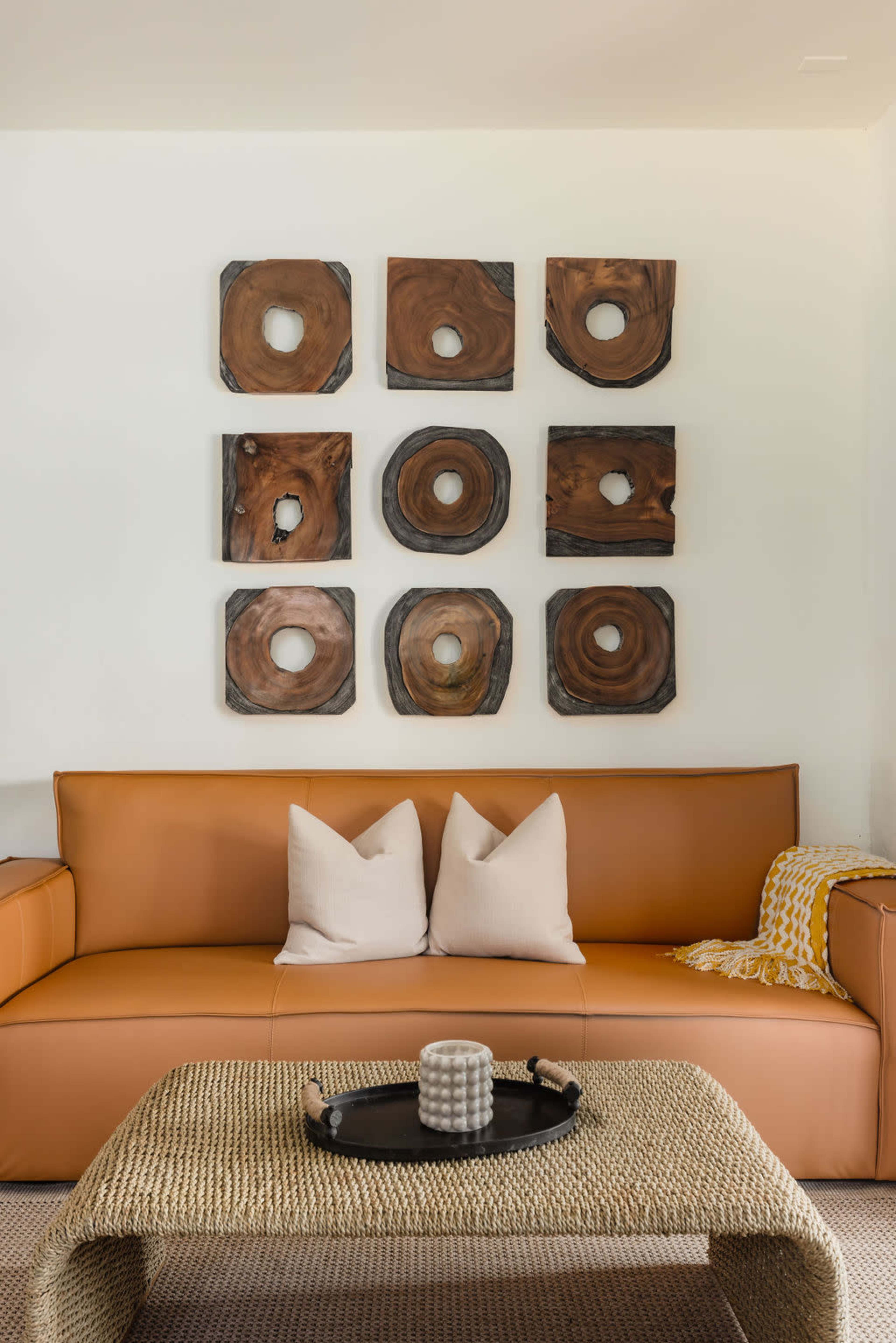 A leather couch with two decorative pillows sits in front of a woven coffee table, beneath a wall adorned with wooden circular art pieces.