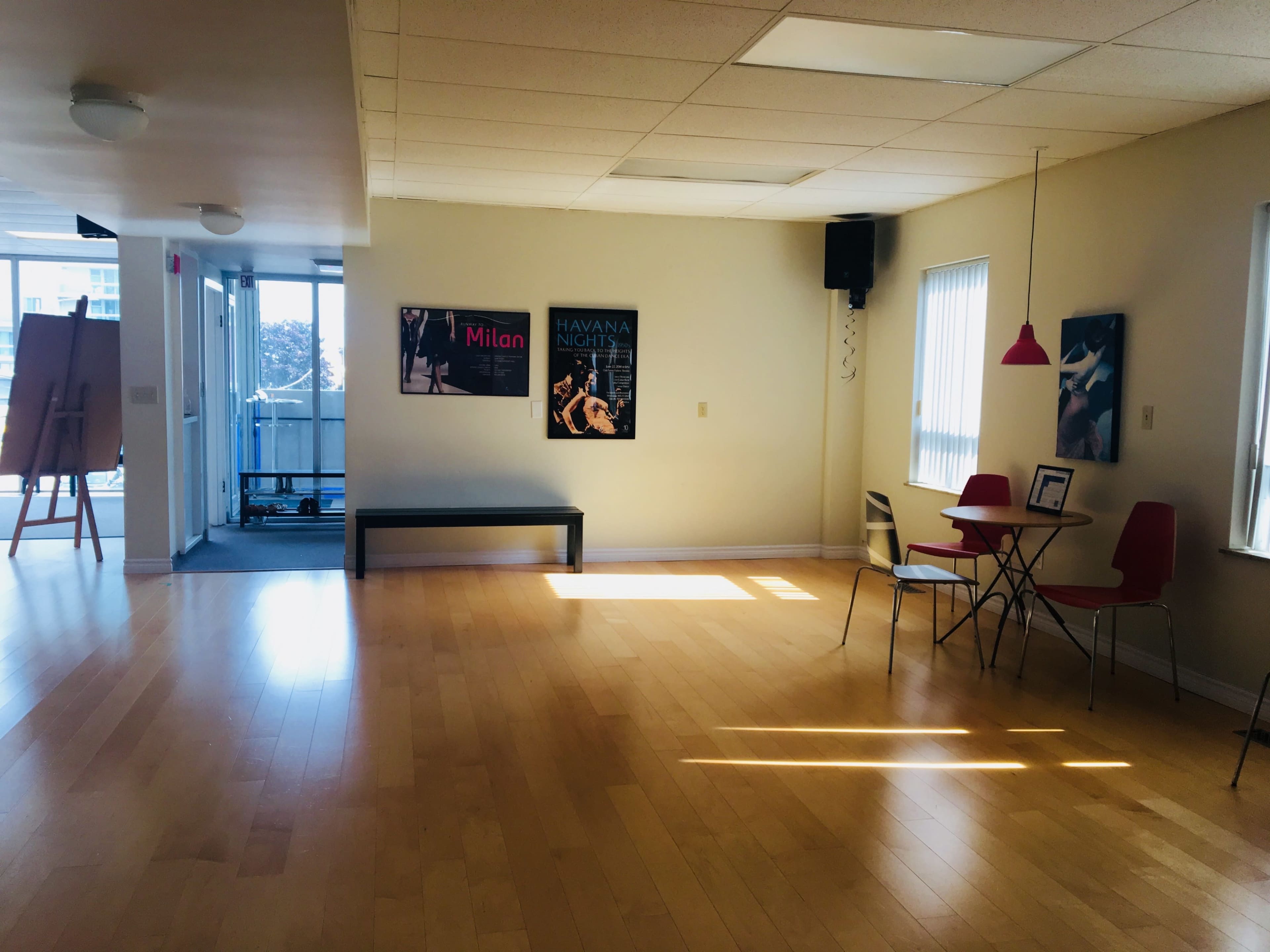 A bright, empty room with wooden flooring, a bench against one wall, a small dining table with red chairs, and posters hanging on the walls.