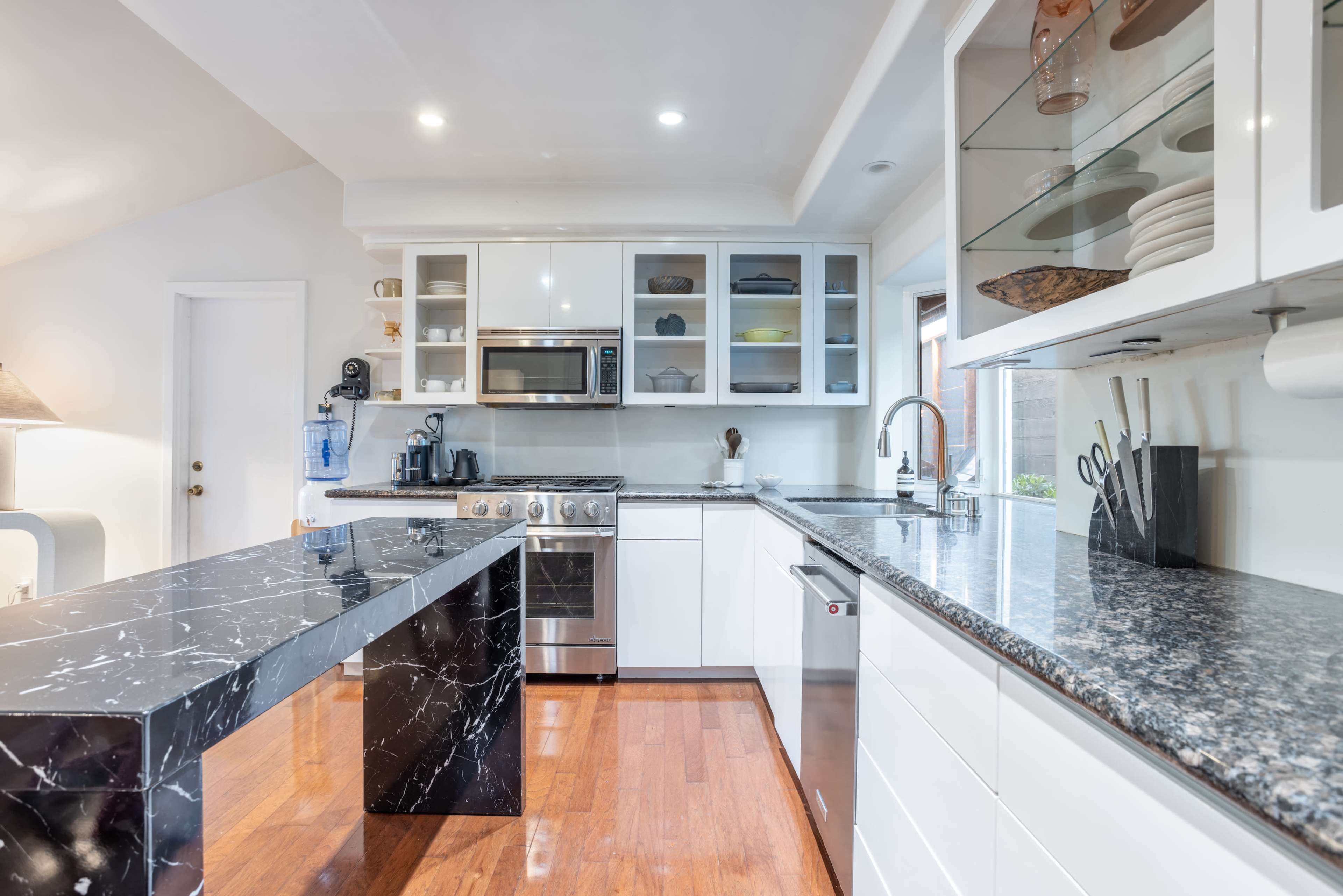 A modern kitchen features sleek white cabinets, a stainless steel stove and microwave, and a black marble island with wooden flooring.