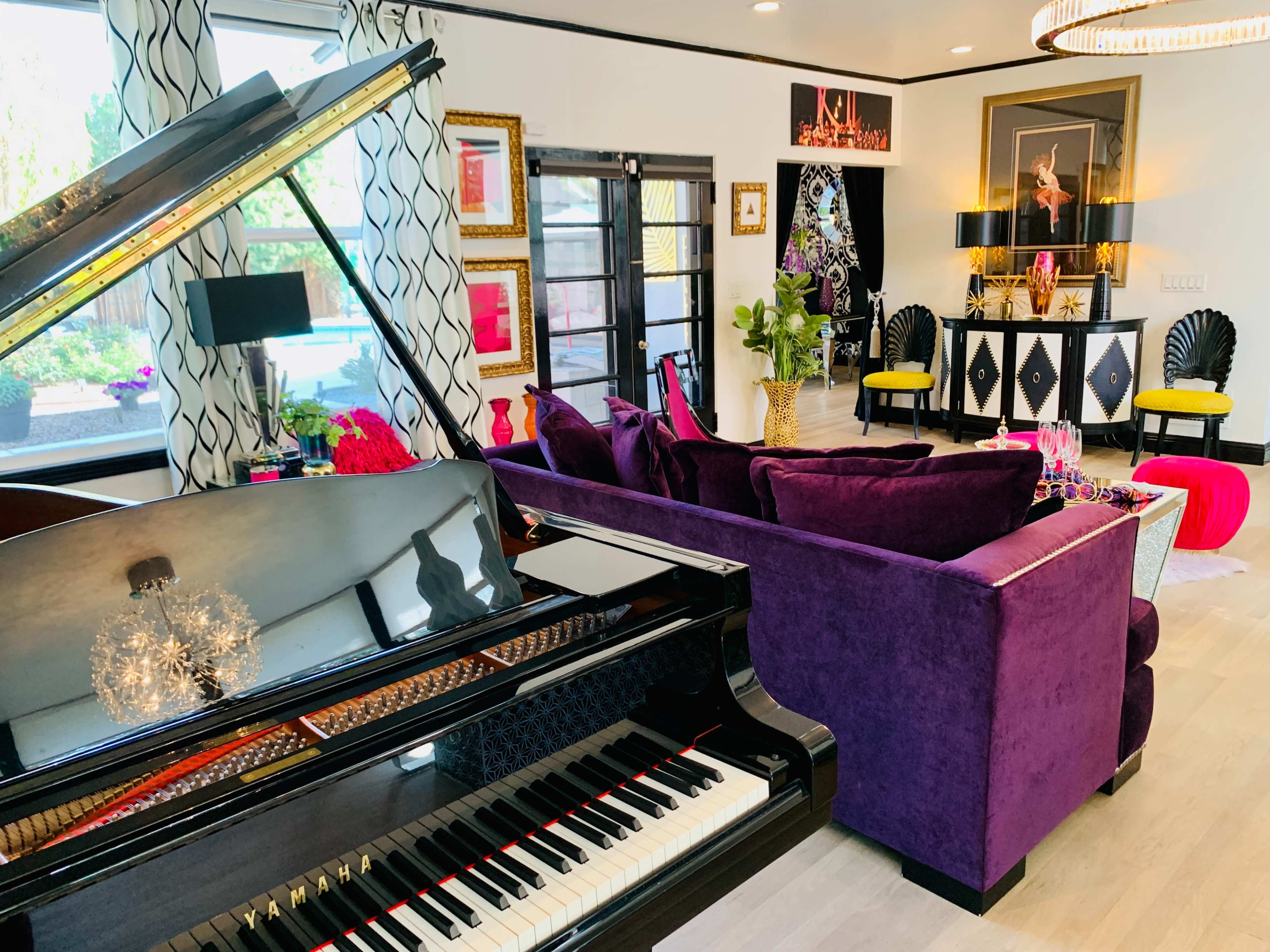 A black grand piano is positioned in the foreground of a modern living room featuring purple couches and vibrant decor.