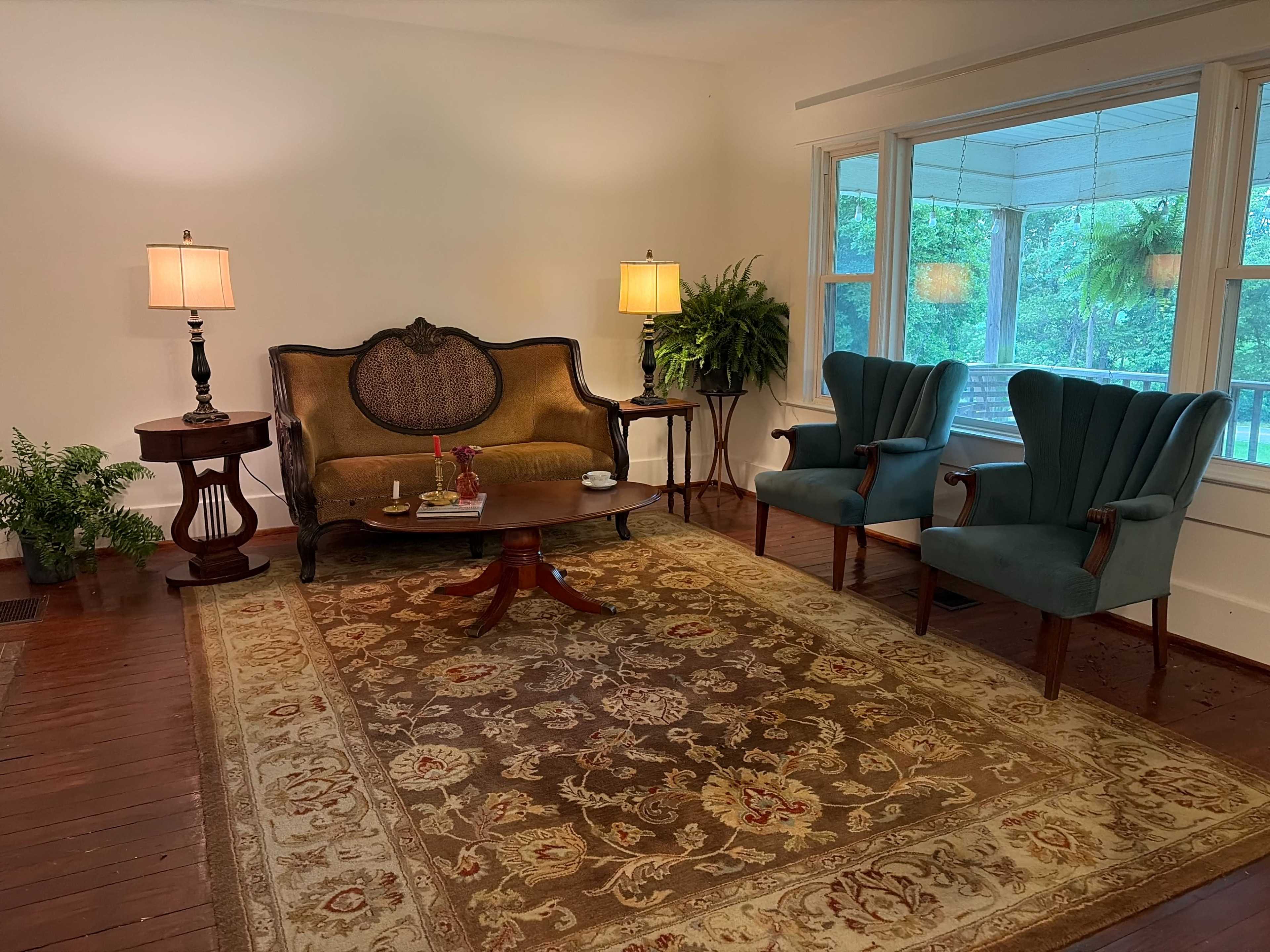 The image shows a cozy living room with a vintage sofa, two blue armchairs, a coffee table, and a large rug, illuminated by two table lamps.