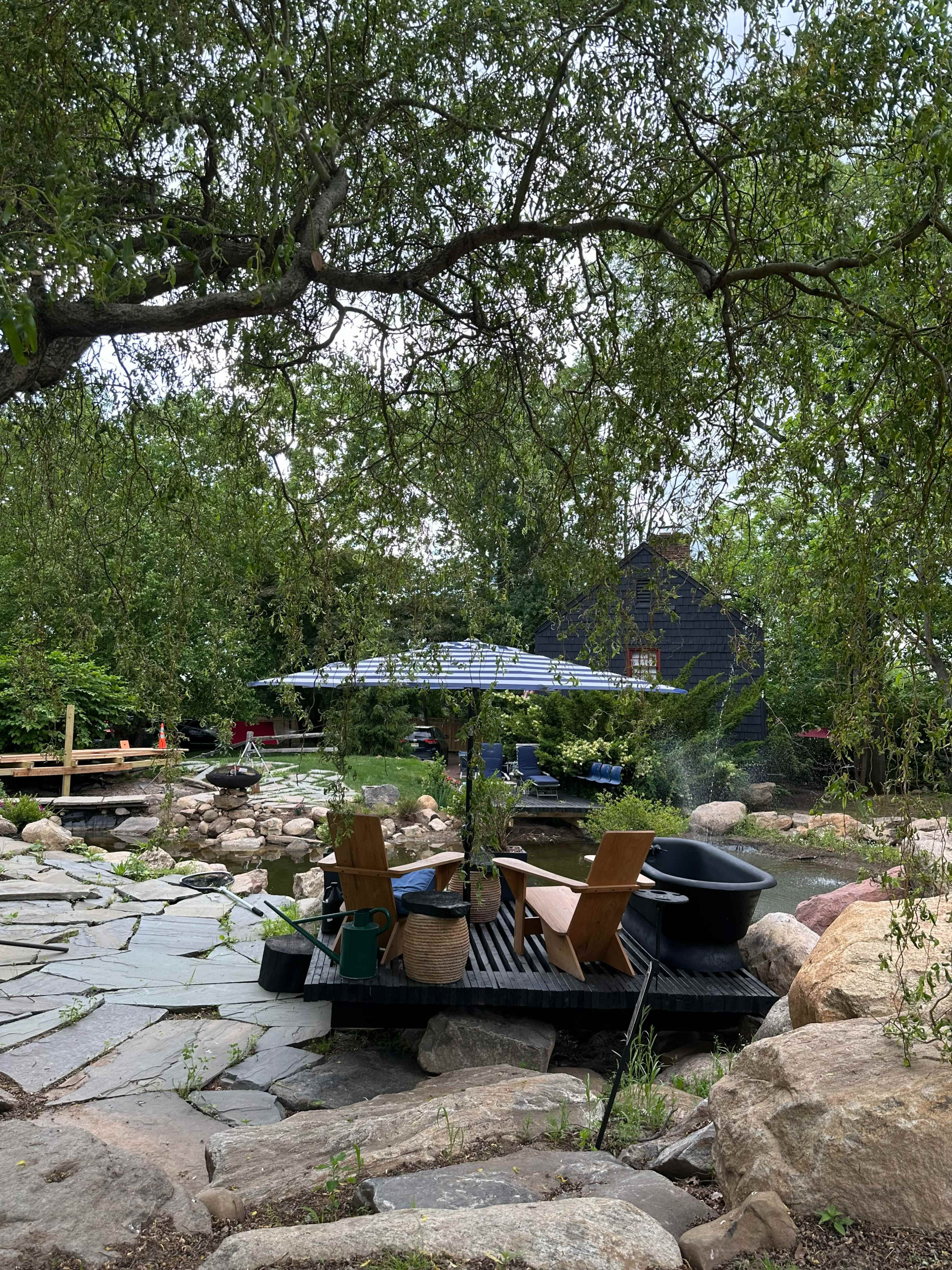 The image shows a stone pathway winding through a garden with wooden chairs, a small table, and an umbrella near a collection of rocks and plants.