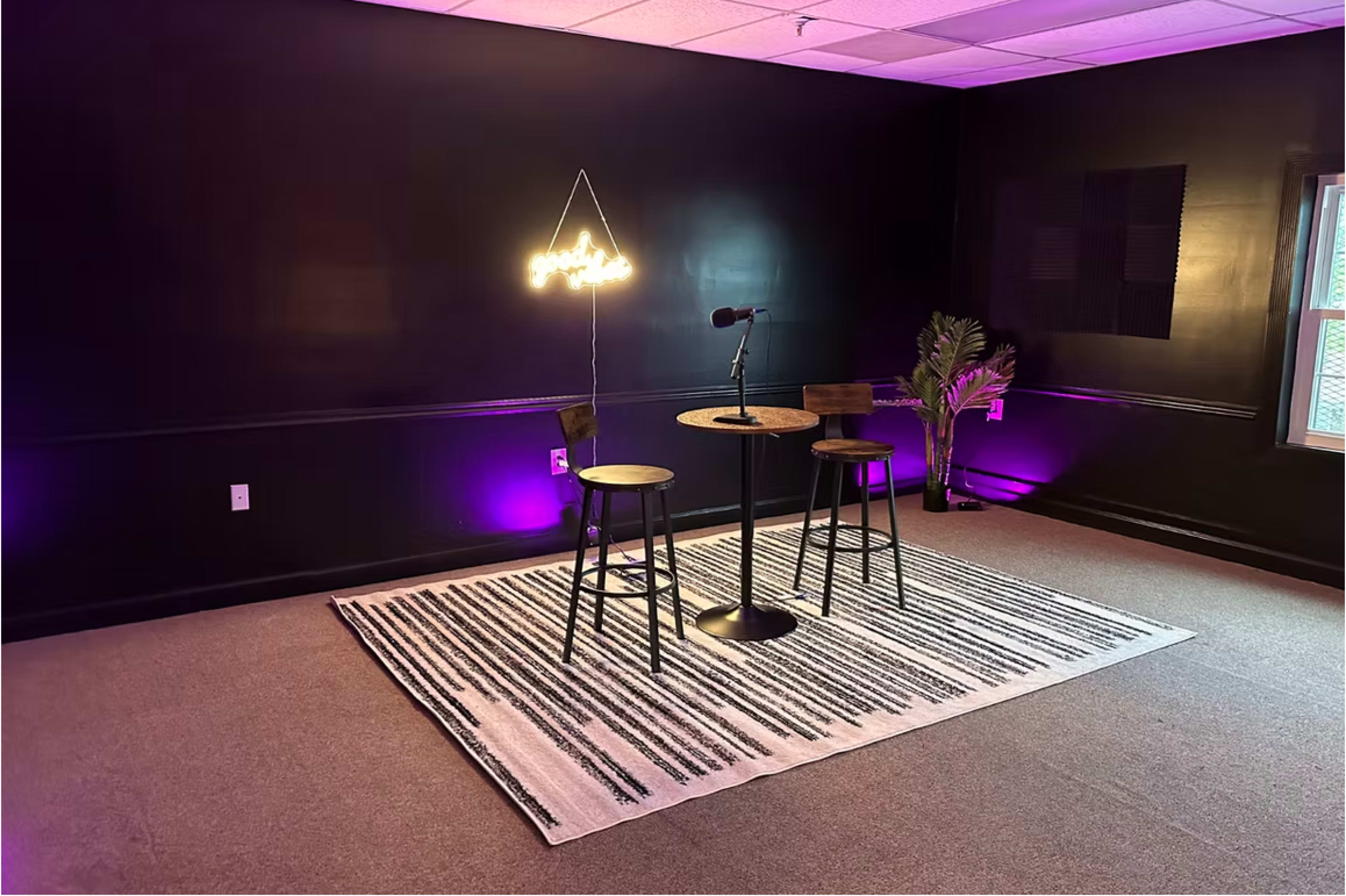 The image shows a small studio space with two bar stools and a round table under a hanging light, surrounded by purple and warm lighting on a patterned rug.