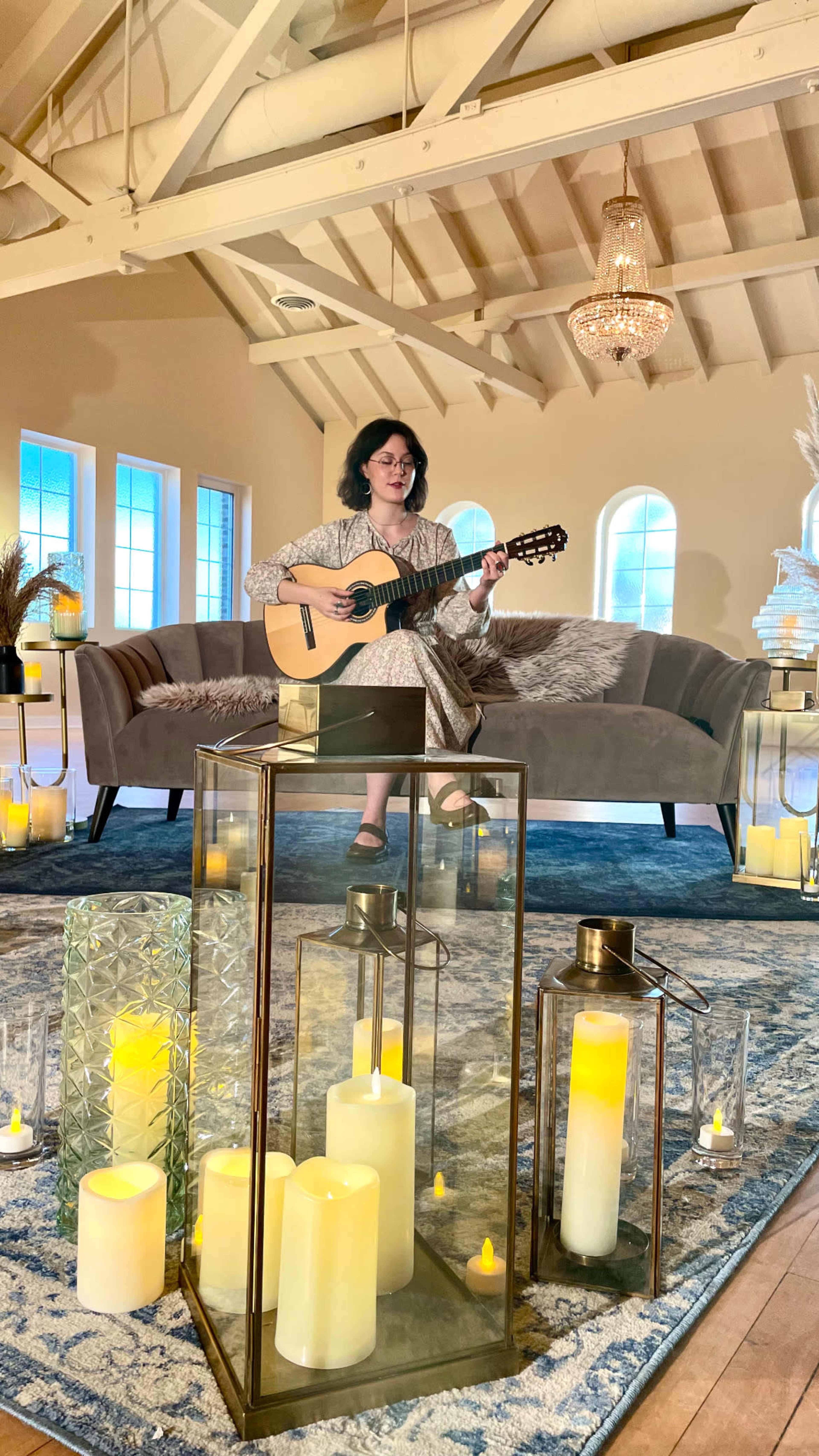 A person sits on a sofa playing guitar, surrounded by decorative lanterns and candles in a softly lit room with large windows.