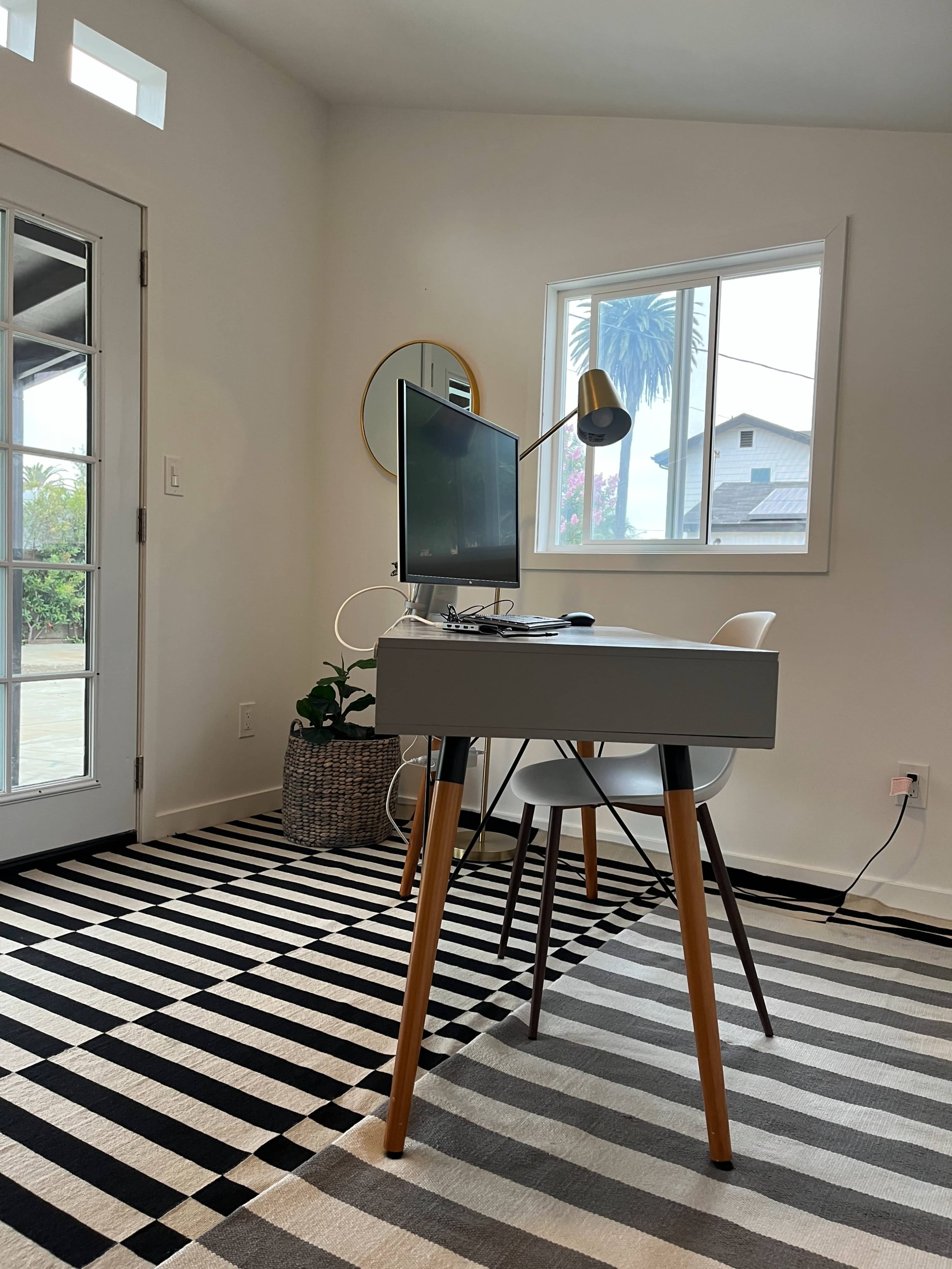 A modern workspace features a desk with a computer and lamp, situated next to a window with a view of greenery.