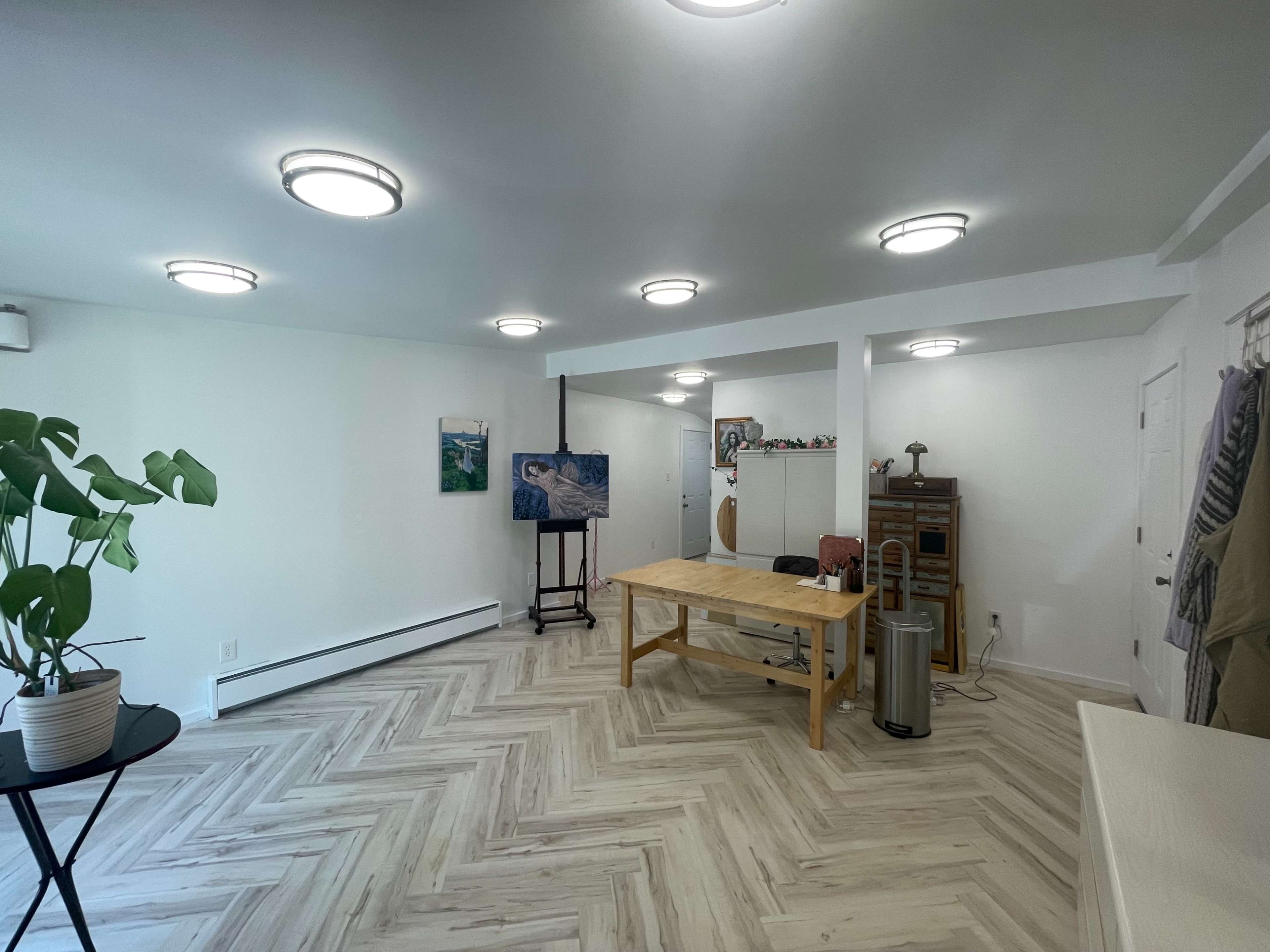 The image shows a spacious, well-lit room with a wooden table, an easel displaying a painting, and several light fixtures on the ceiling.