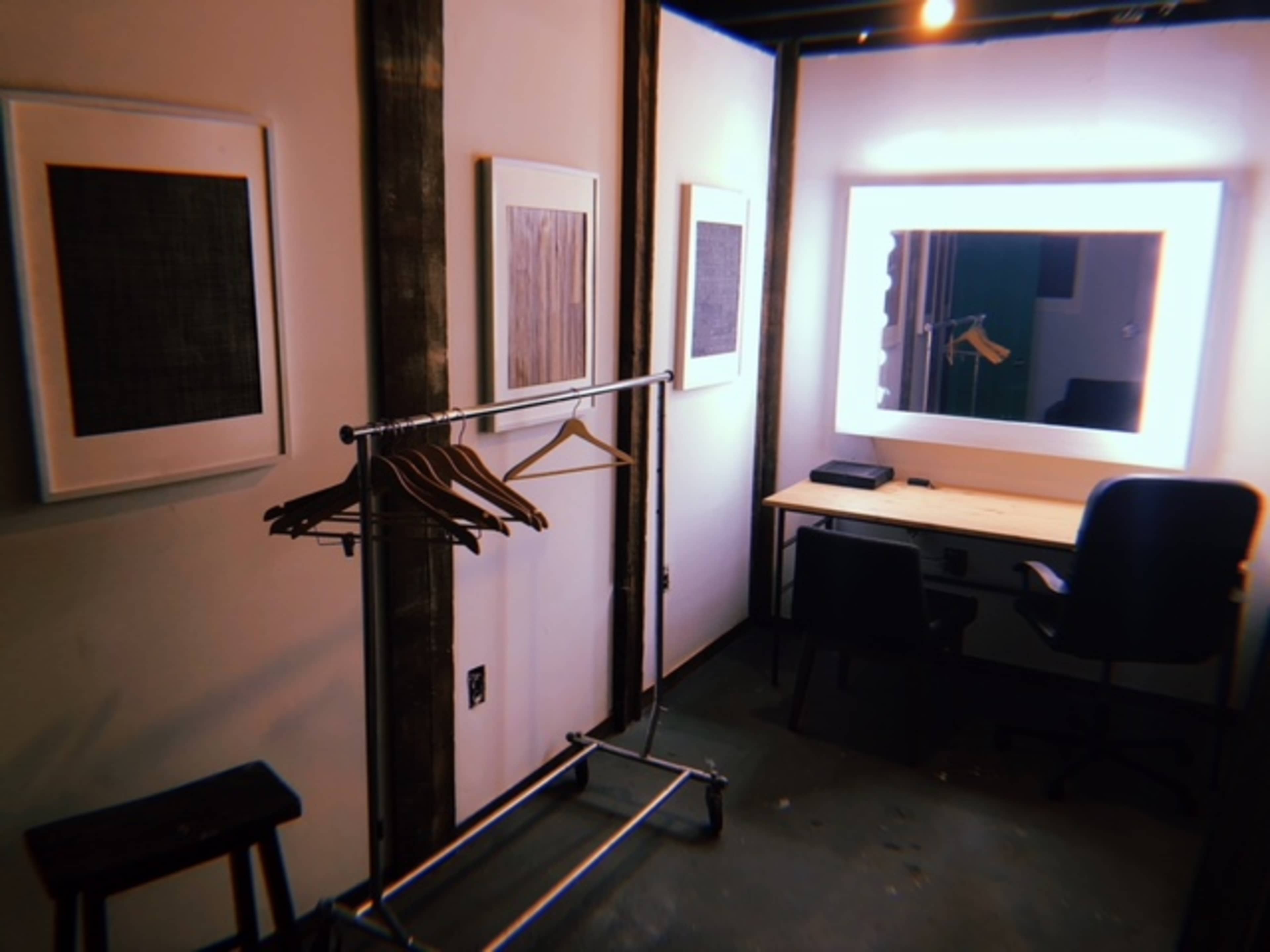 The image shows a minimalist dressing room with clothes hangers on a rack, a desk, a chair, and framed artwork on the walls.