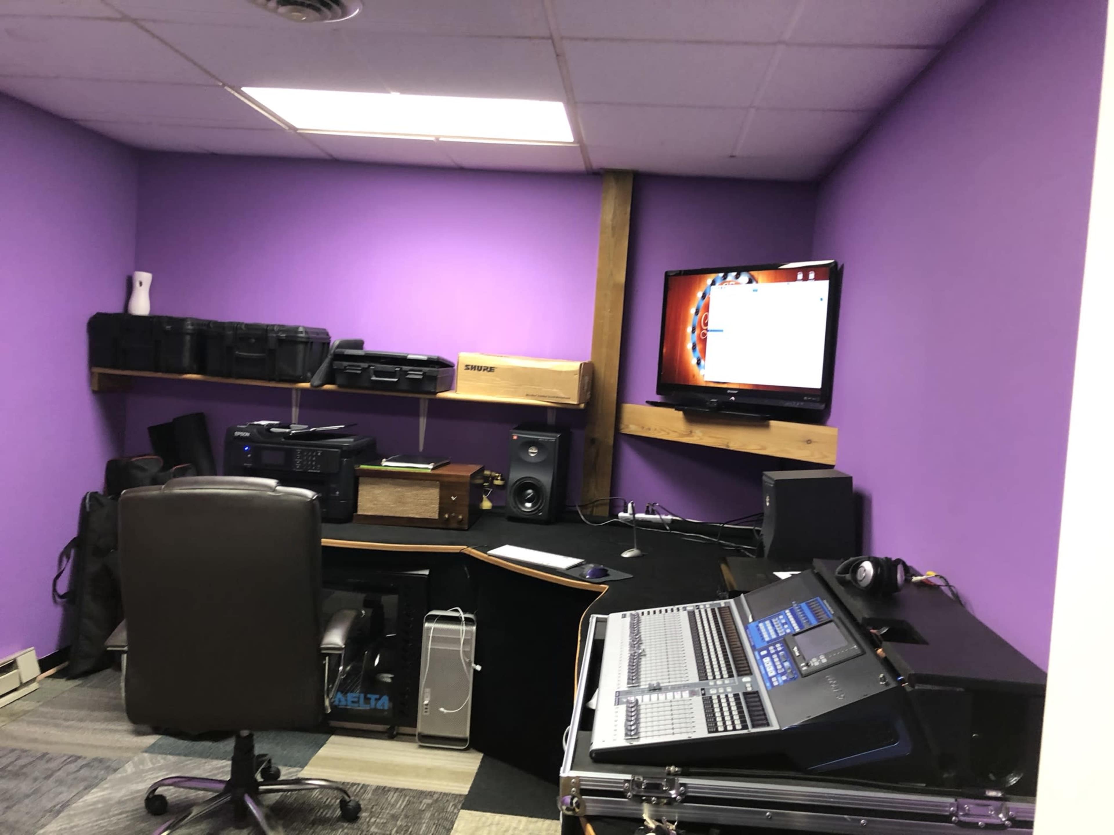 A small recording studio features a purple wall, a computer monitor, audio equipment, and a black office chair positioned at a desk.