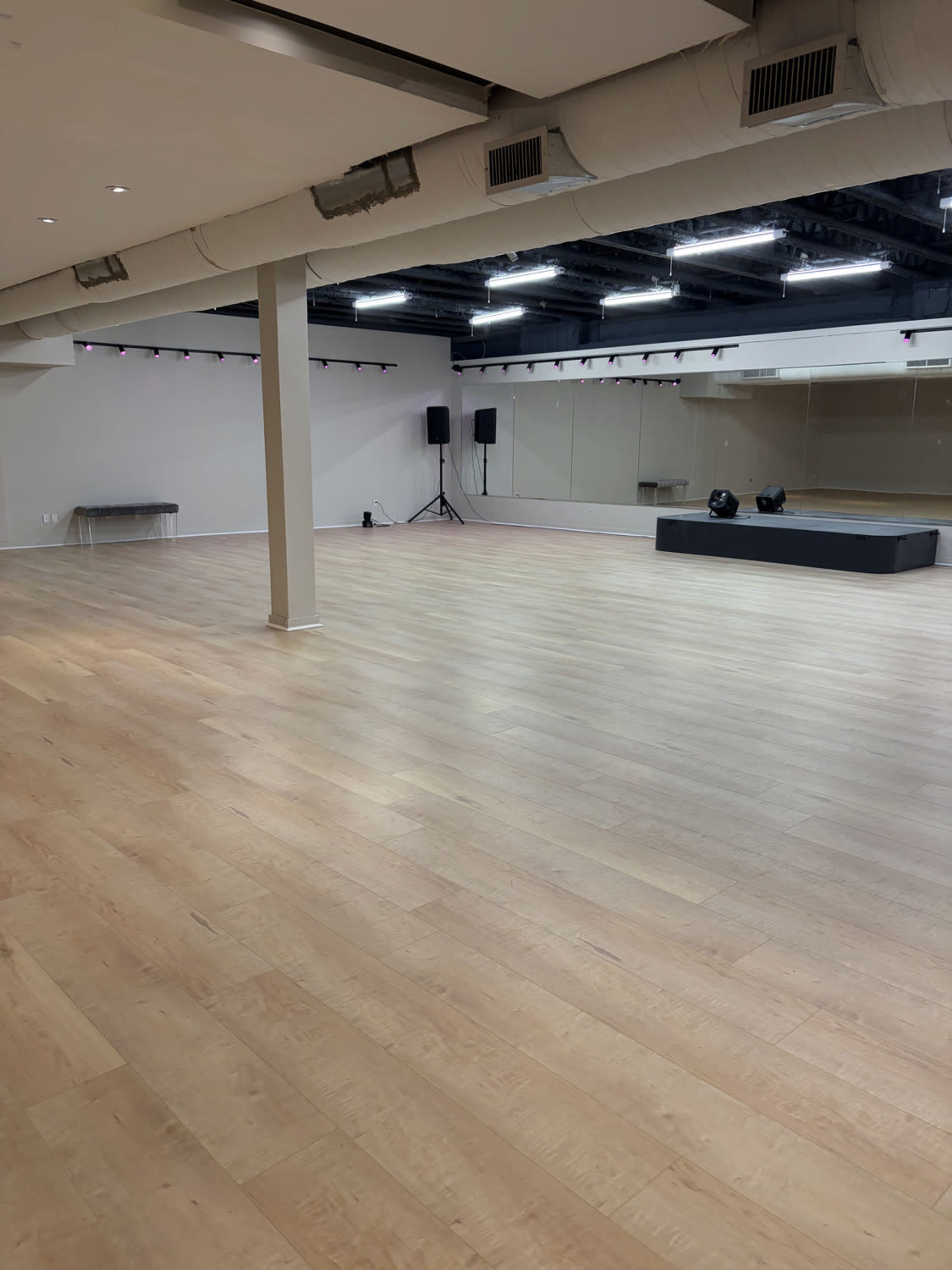 The image shows a spacious, empty room with wooden flooring, equipped with mirrors, a stage area, and sound equipment.