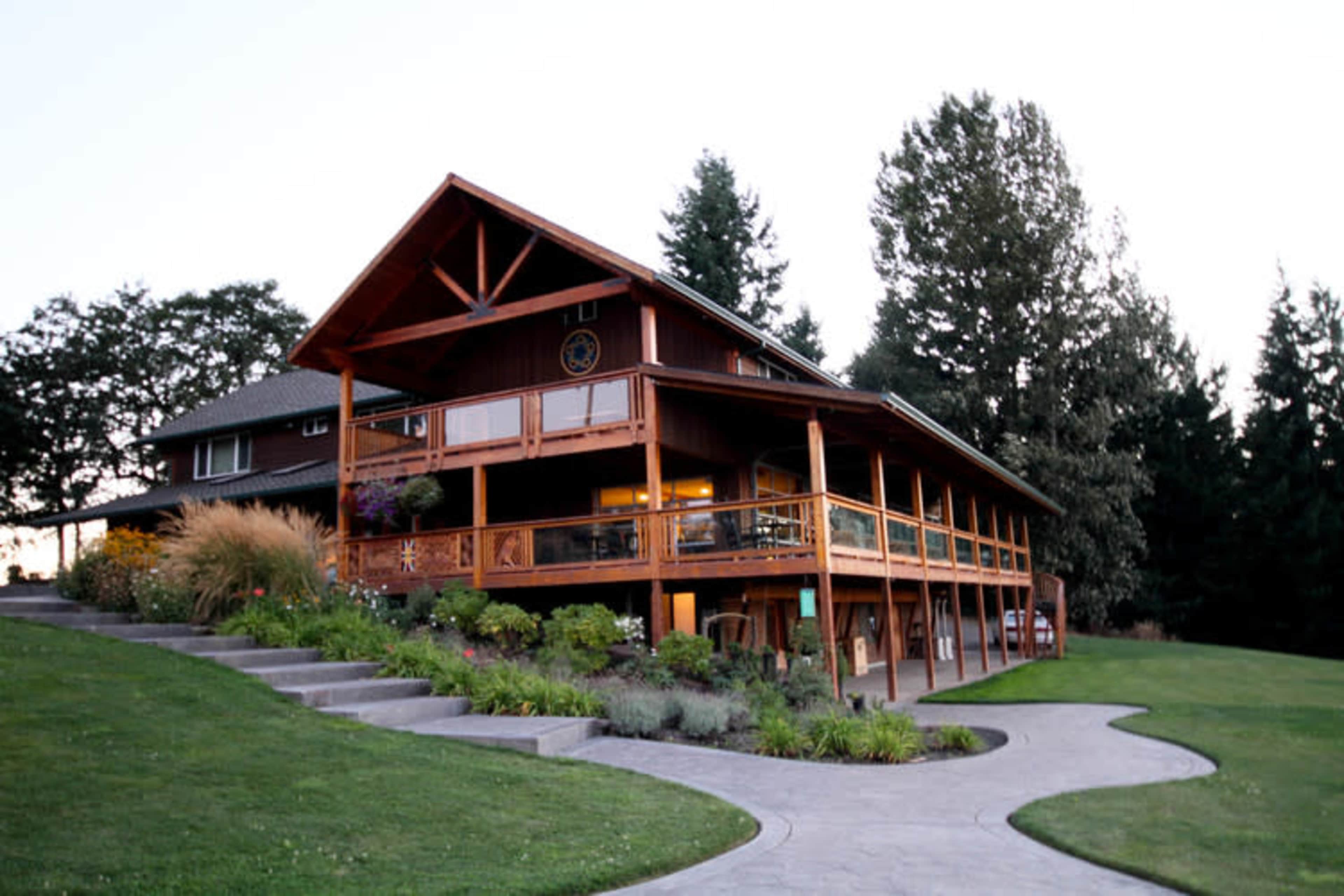 A large wooden house with a landscaped yard and a sloped driveway is set against a backdrop of trees at dusk.
