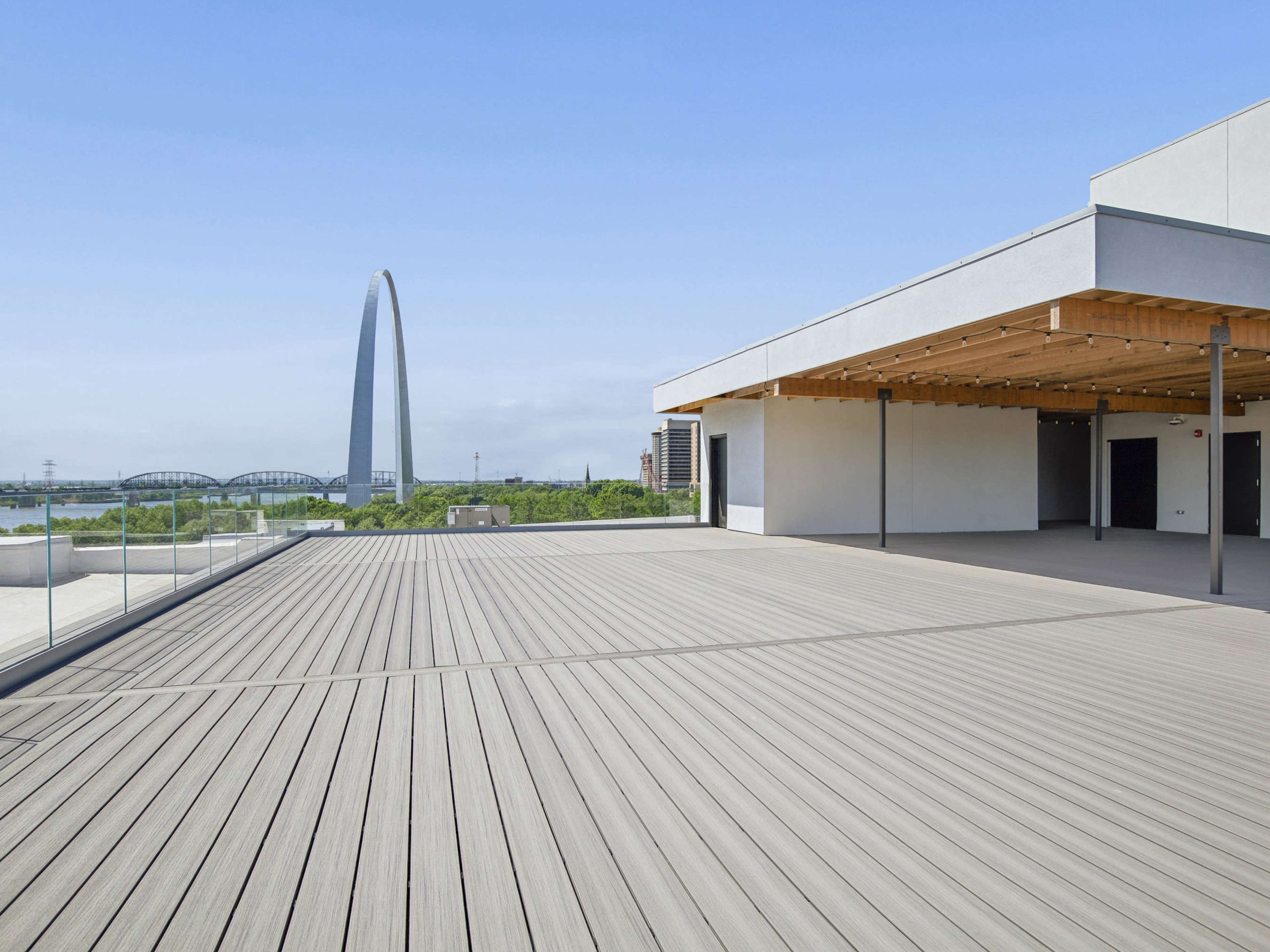 A spacious rooftop deck with wooden flooring overlooks a large arch structure and a river in the distance.