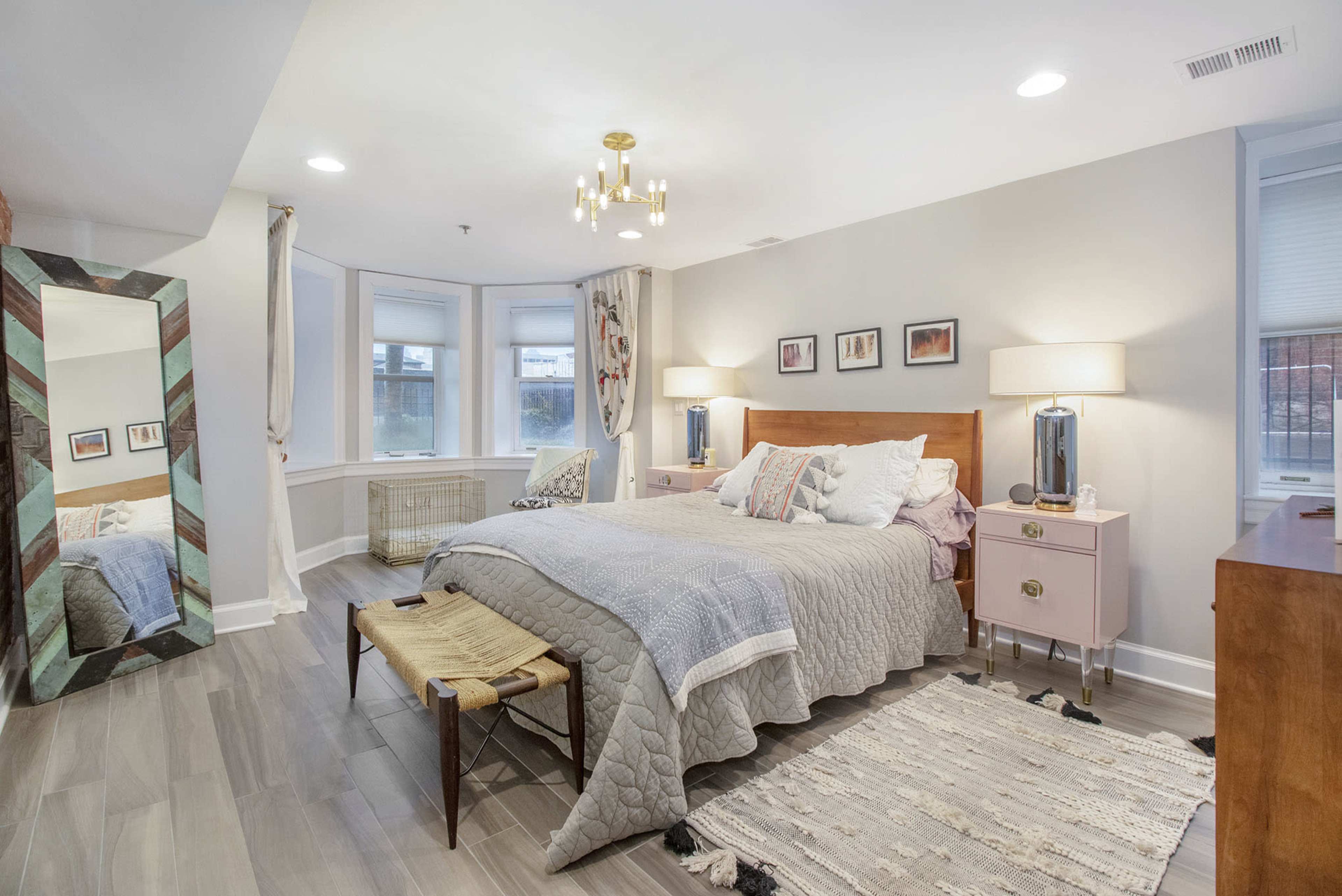 A well-lit bedroom featuring a large bed with gray bedding, a decorative mirror, bedside lamps, and a wooden dresser.