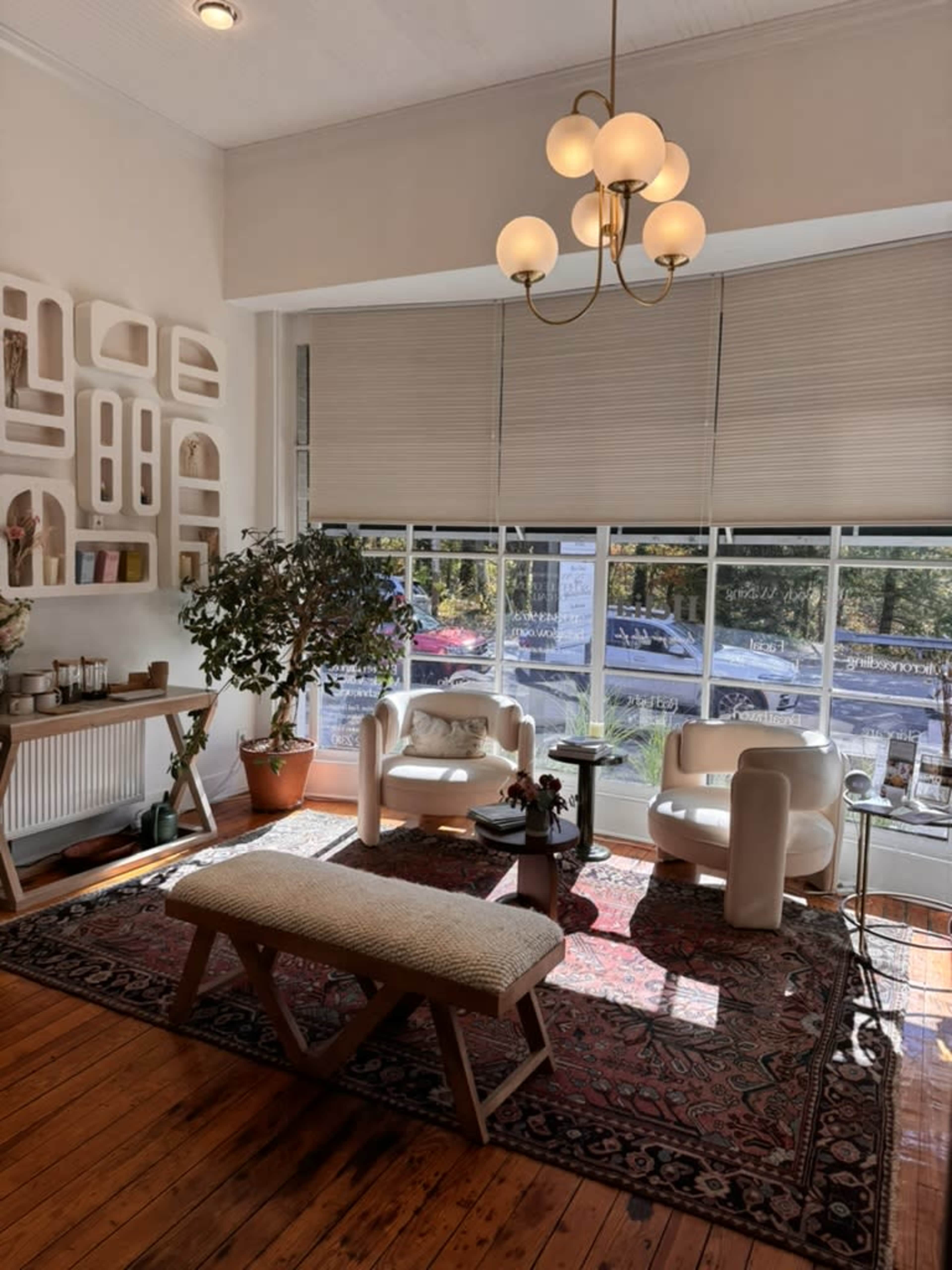 The bright interior features two stylish armchairs, a wooden bench, a coffee table, and large windows that allow natural light to illuminate the space.