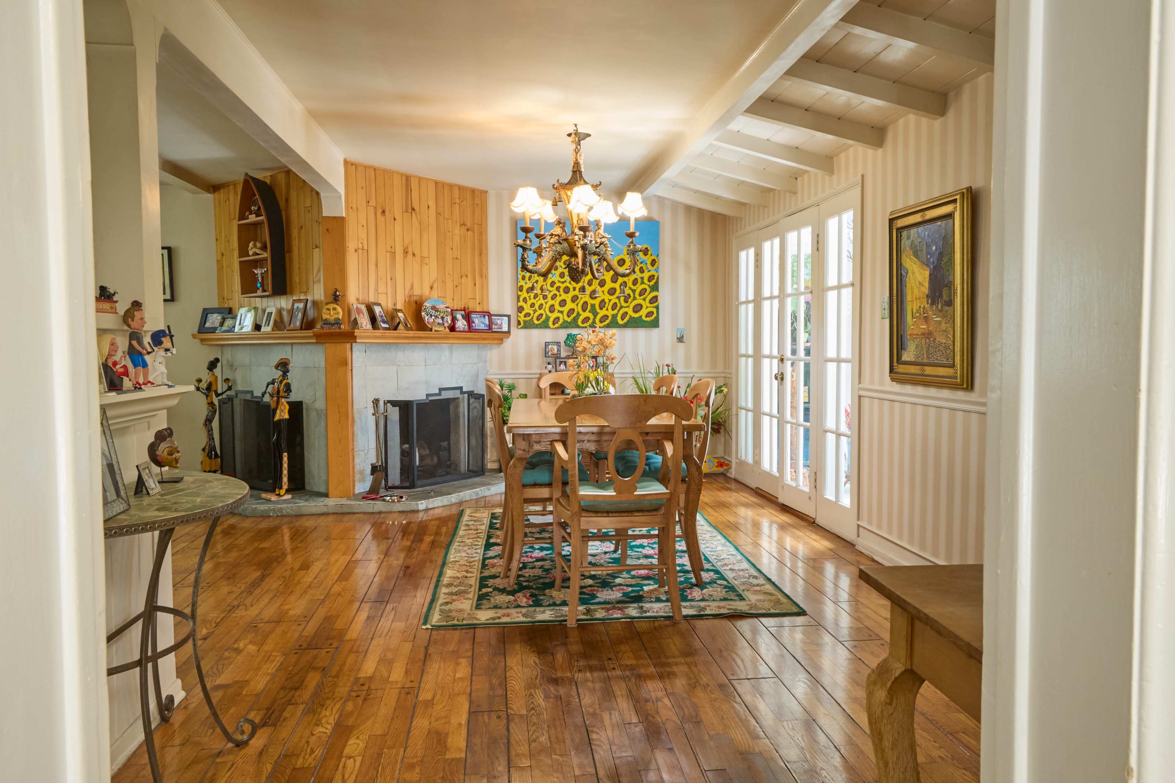 The image shows a brightly lit living room and dining area with wooden floors, a fireplace, and a dining table surrounded by chairs.