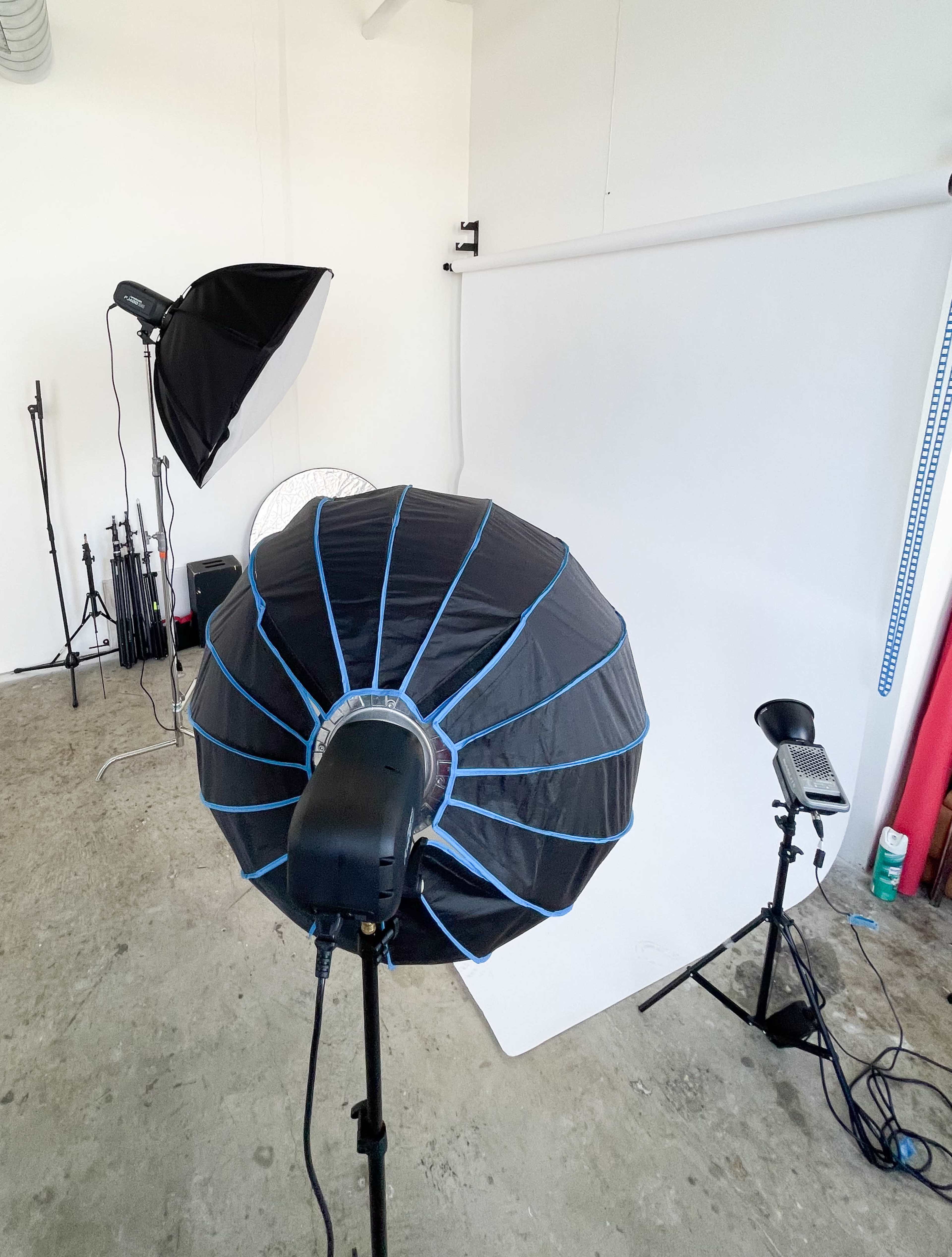 The image shows a photography studio setup with a large softbox light and various lighting equipment against a white backdrop.