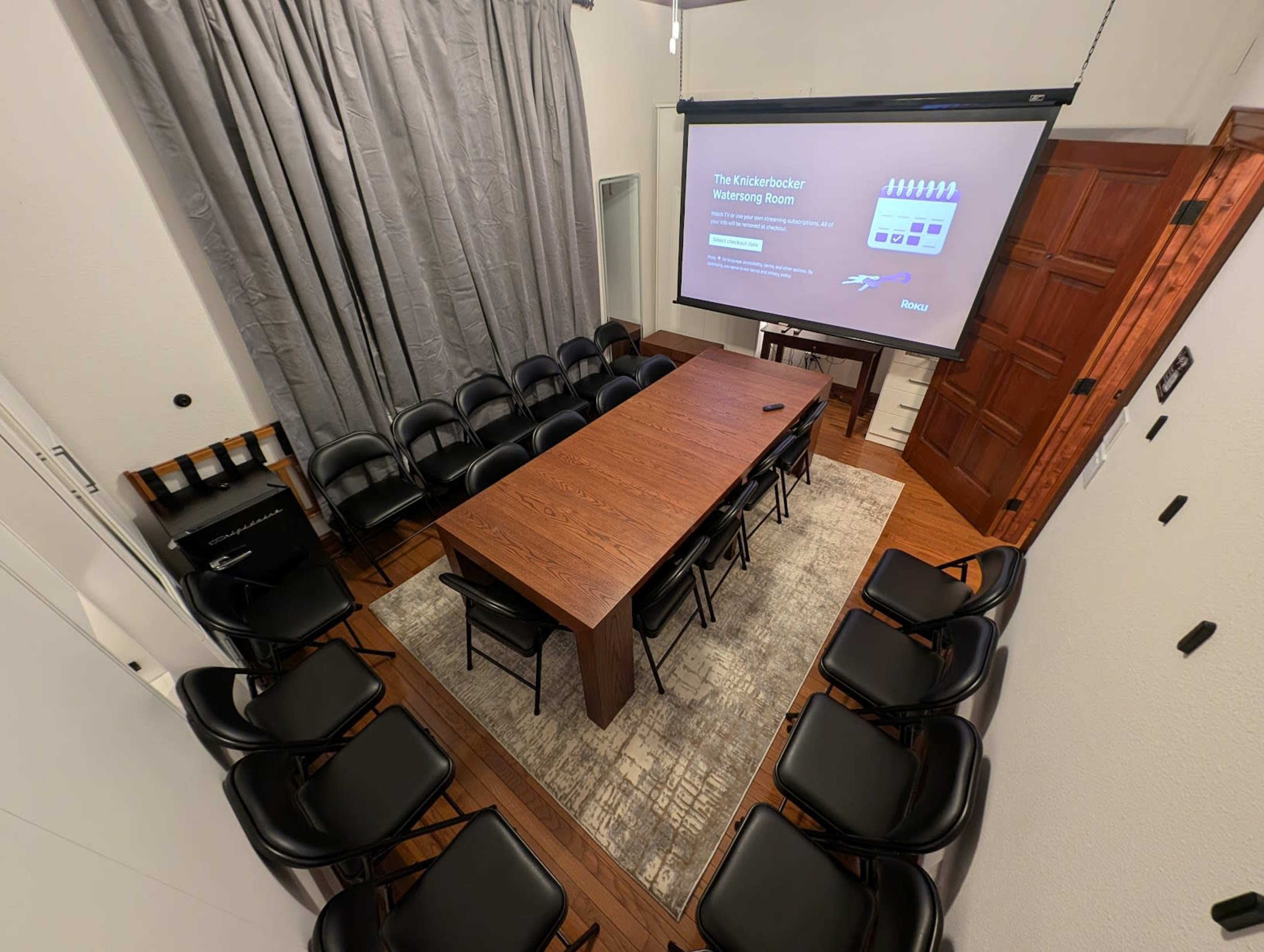 The image shows a rectangular conference room with a long wooden table surrounded by black chairs, a projector screen displaying information on one wall, and gray curtains on another wall.