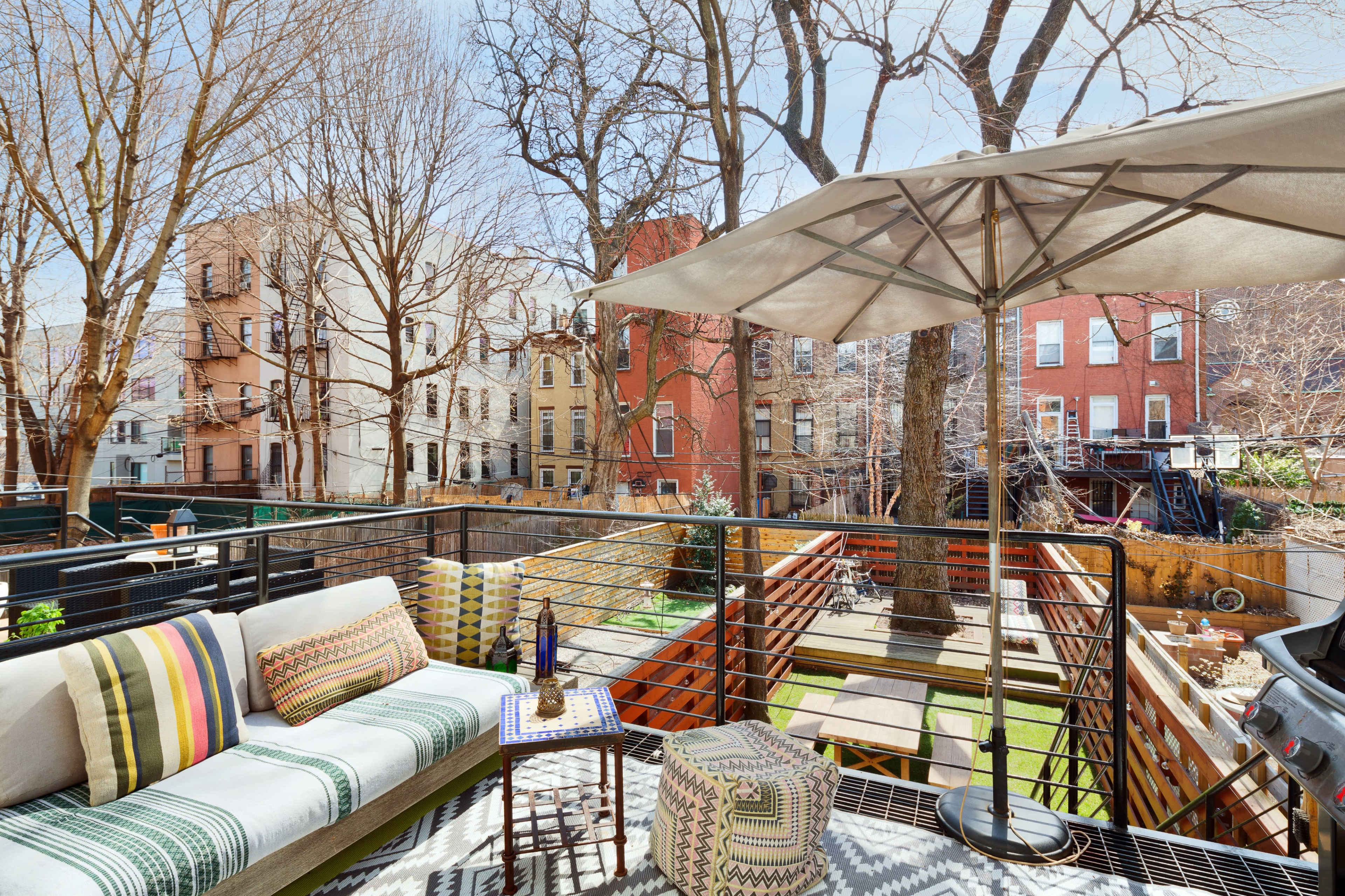 A terrace features a seating area with a sofa, umbrella, and small table overlooking a backyard with trees and buildings in the background.