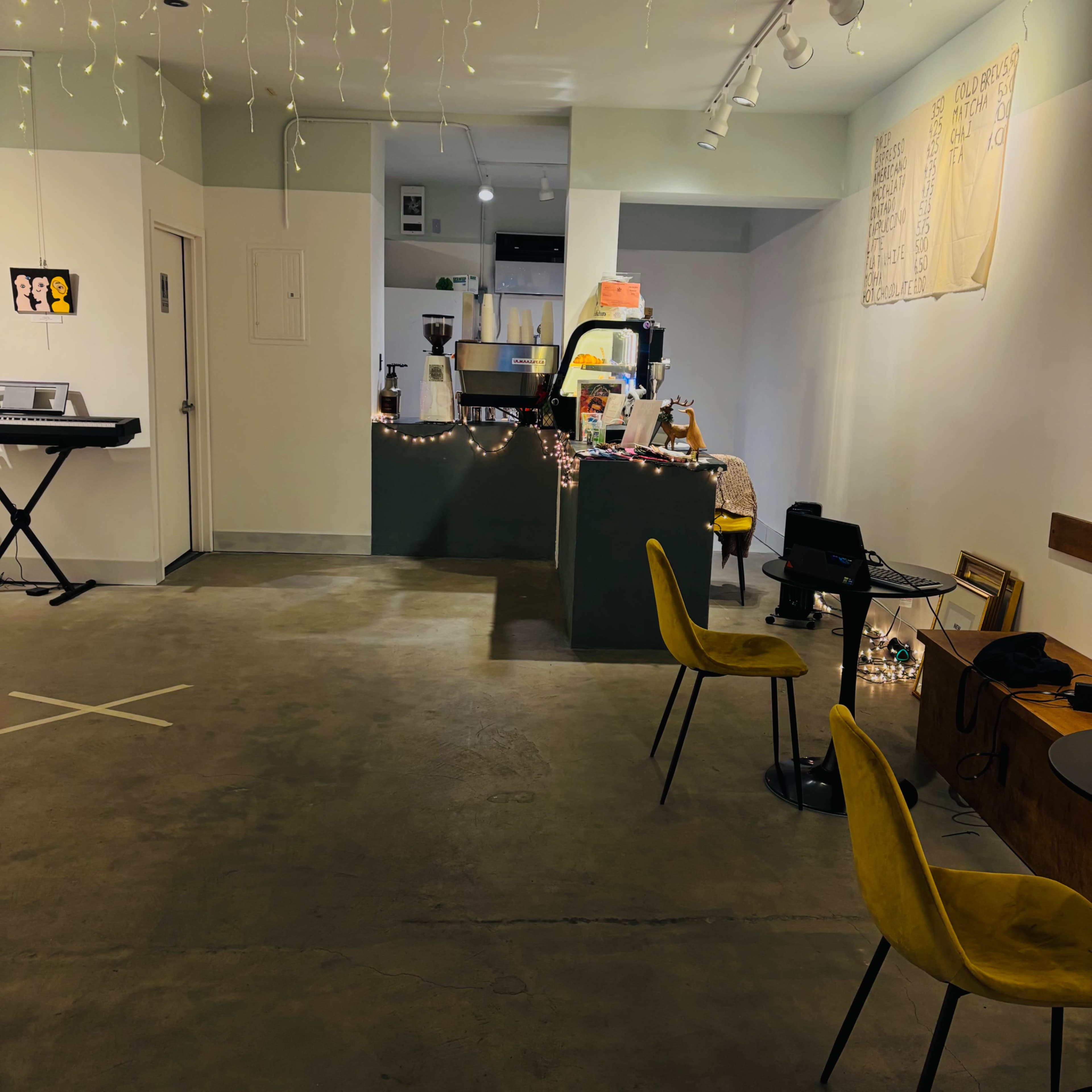 A brightly lit café features a service counter, seating areas with yellow chairs, and decorative string lights hanging from the ceiling.