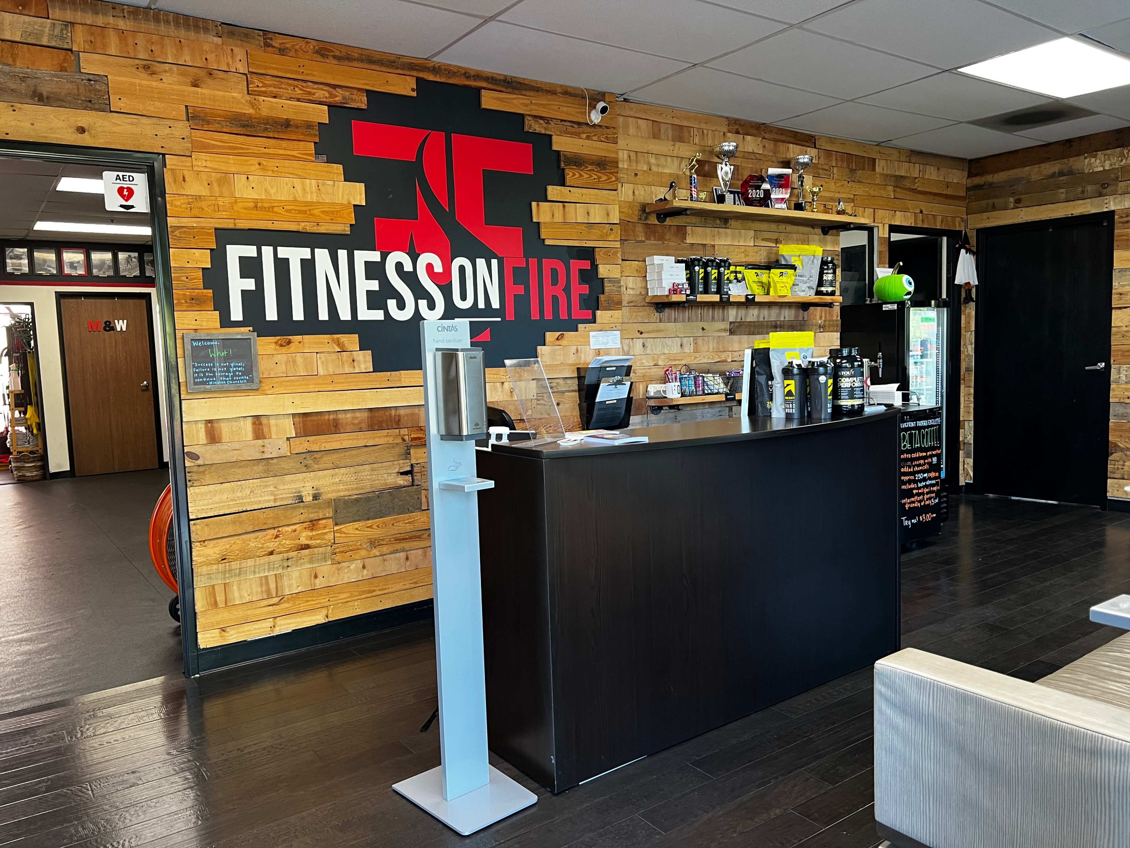 The interior of a fitness center features a reception desk with a backdrop of wooden planks and a large wall graphic that reads "FITNESS ON FIRE."