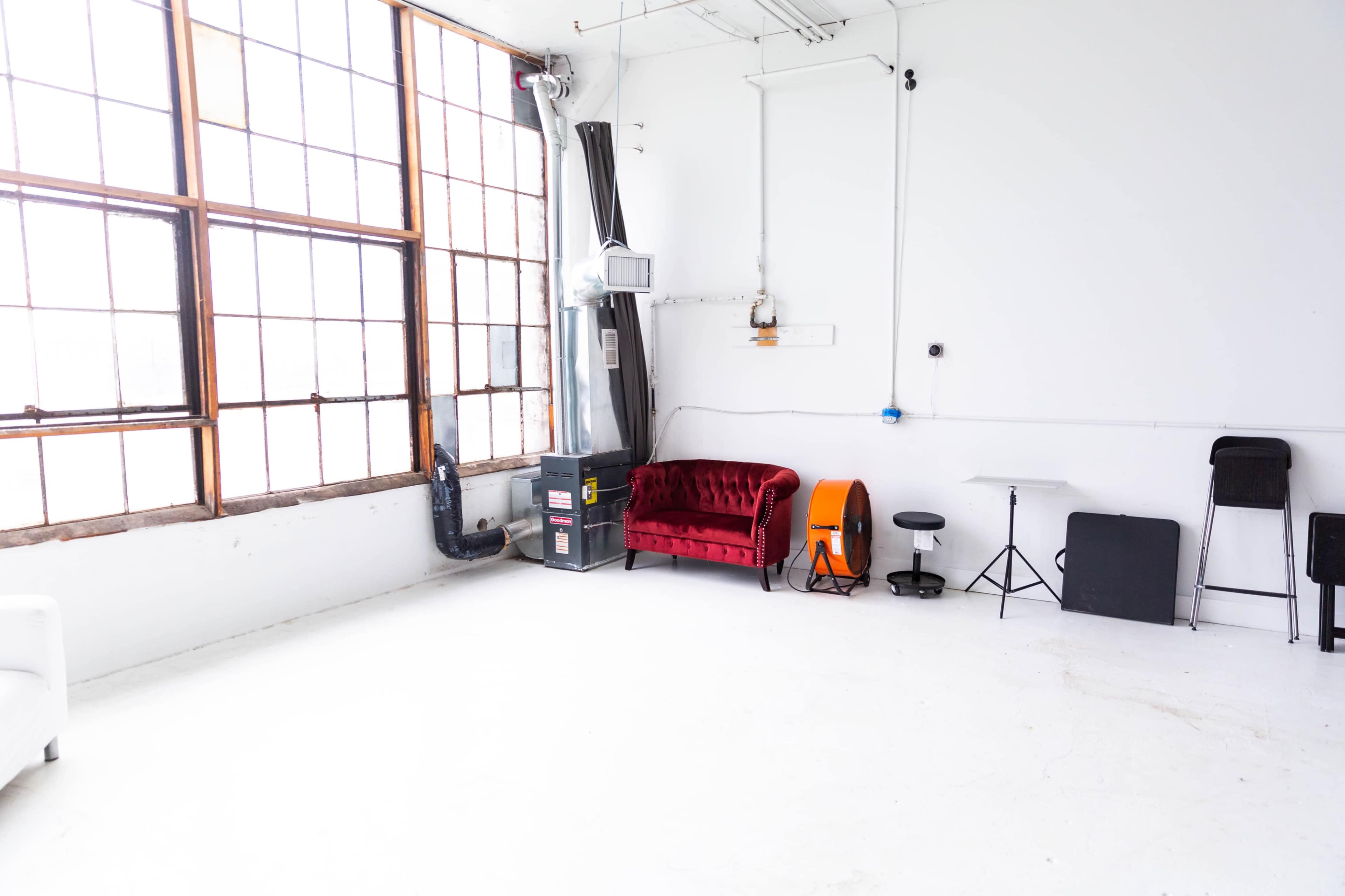 The image shows a bright, empty studio space featuring a red velvet couch, an orange stool, and several black folding chairs against white walls and large windows.