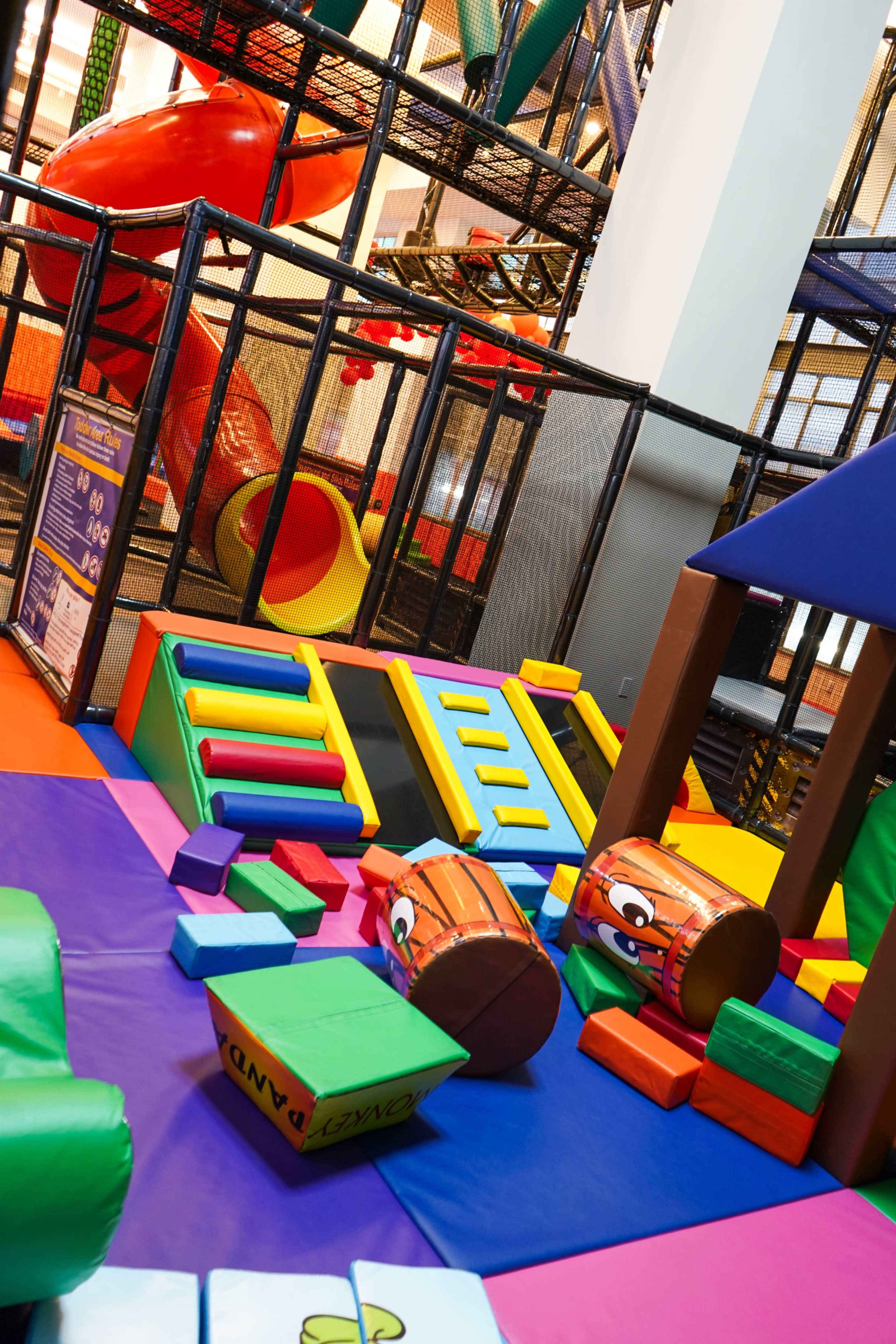 A colorful indoor playground features various climbing structures, slides, and soft play materials scattered across the floor.