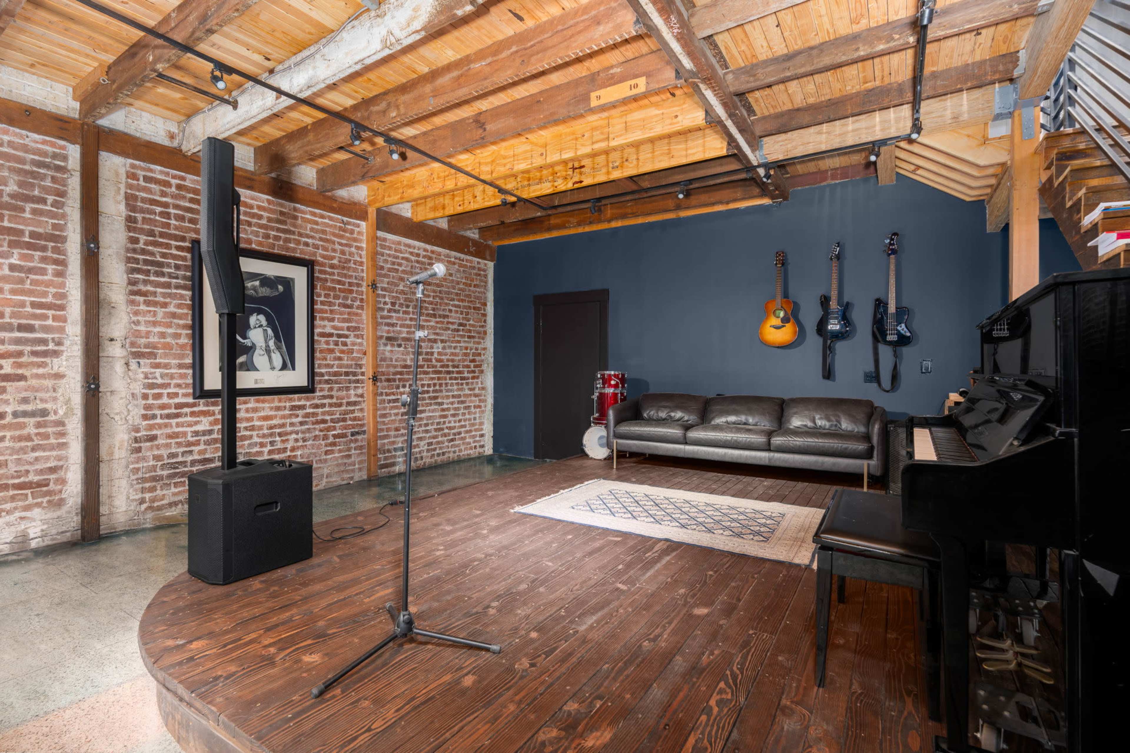 A music studio featuring a wooden platform with a microphone stand, a black piano, and various musical instruments displayed on a dark blue wall, all set within a rustic space with exposed brick and wooden beams.