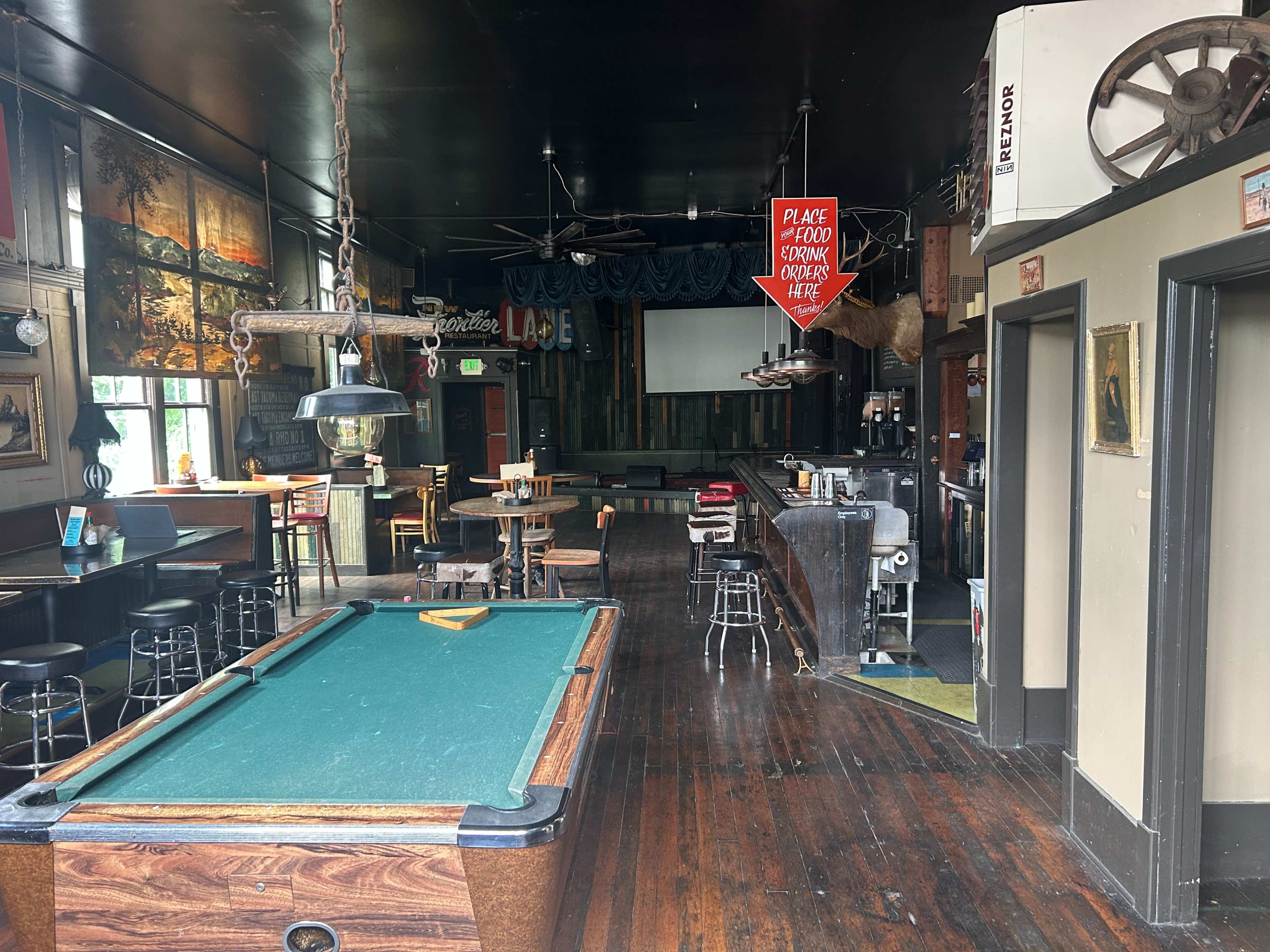 The image shows a bar interior with a billiards table, seating areas, and a projector screen at the far end.