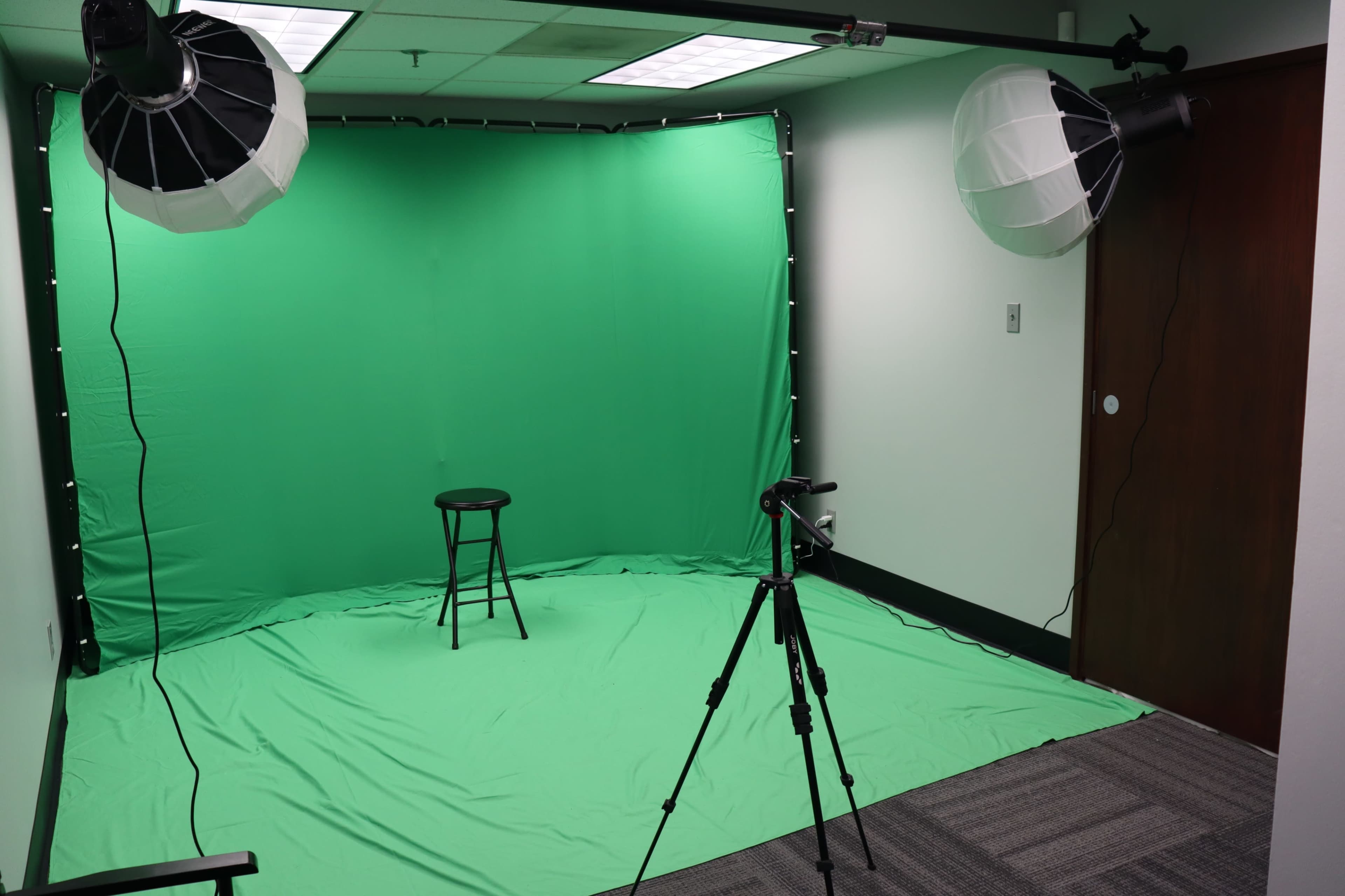 The image shows a small studio setup with a green backdrop, a black stool placed in front, and softbox lights mounted on stands, along with a tripod for a camera.