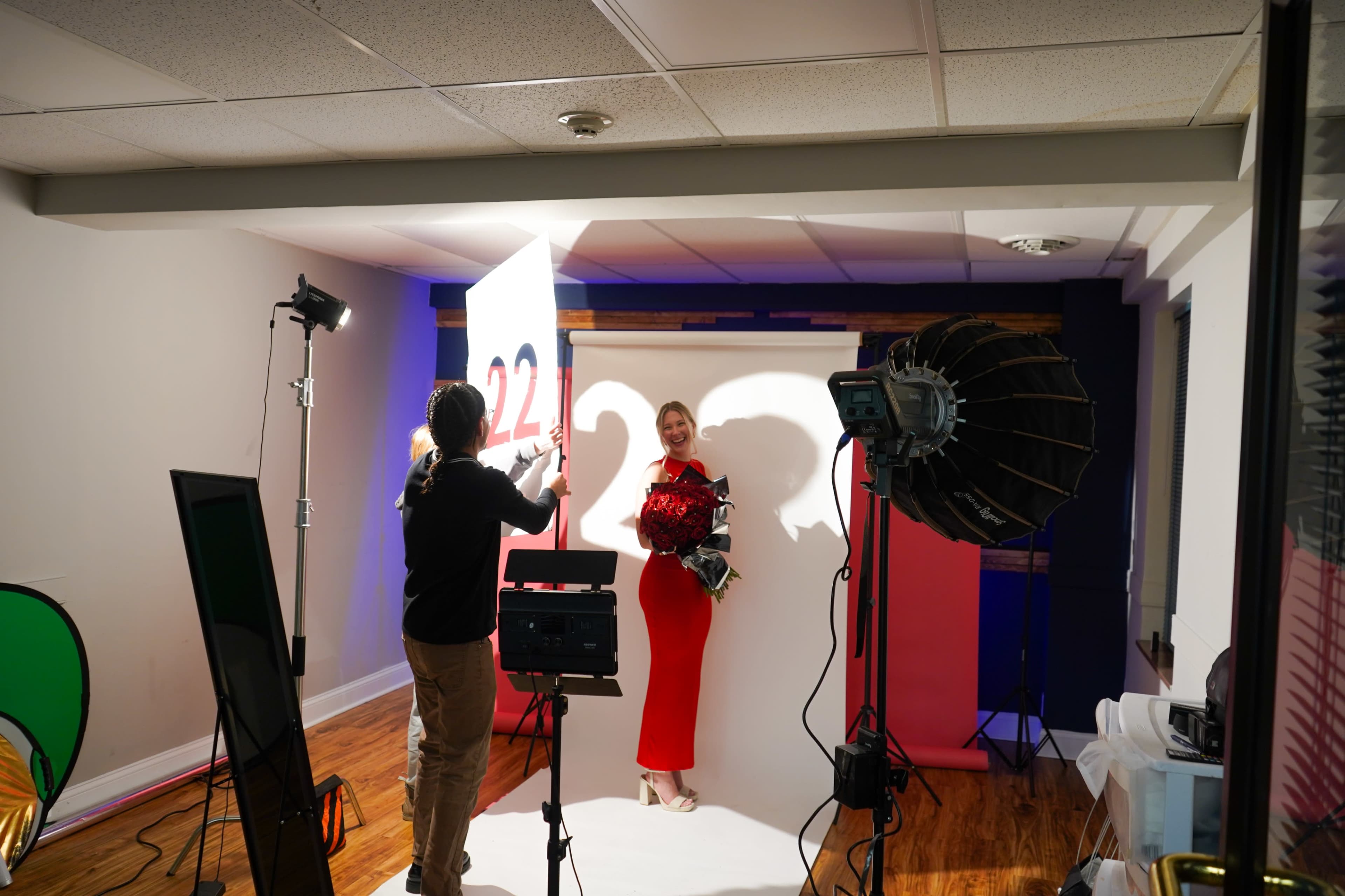 A model in a red dress poses with a bouquet in a studio setting, while a photographer adjusts lighting and equipment.