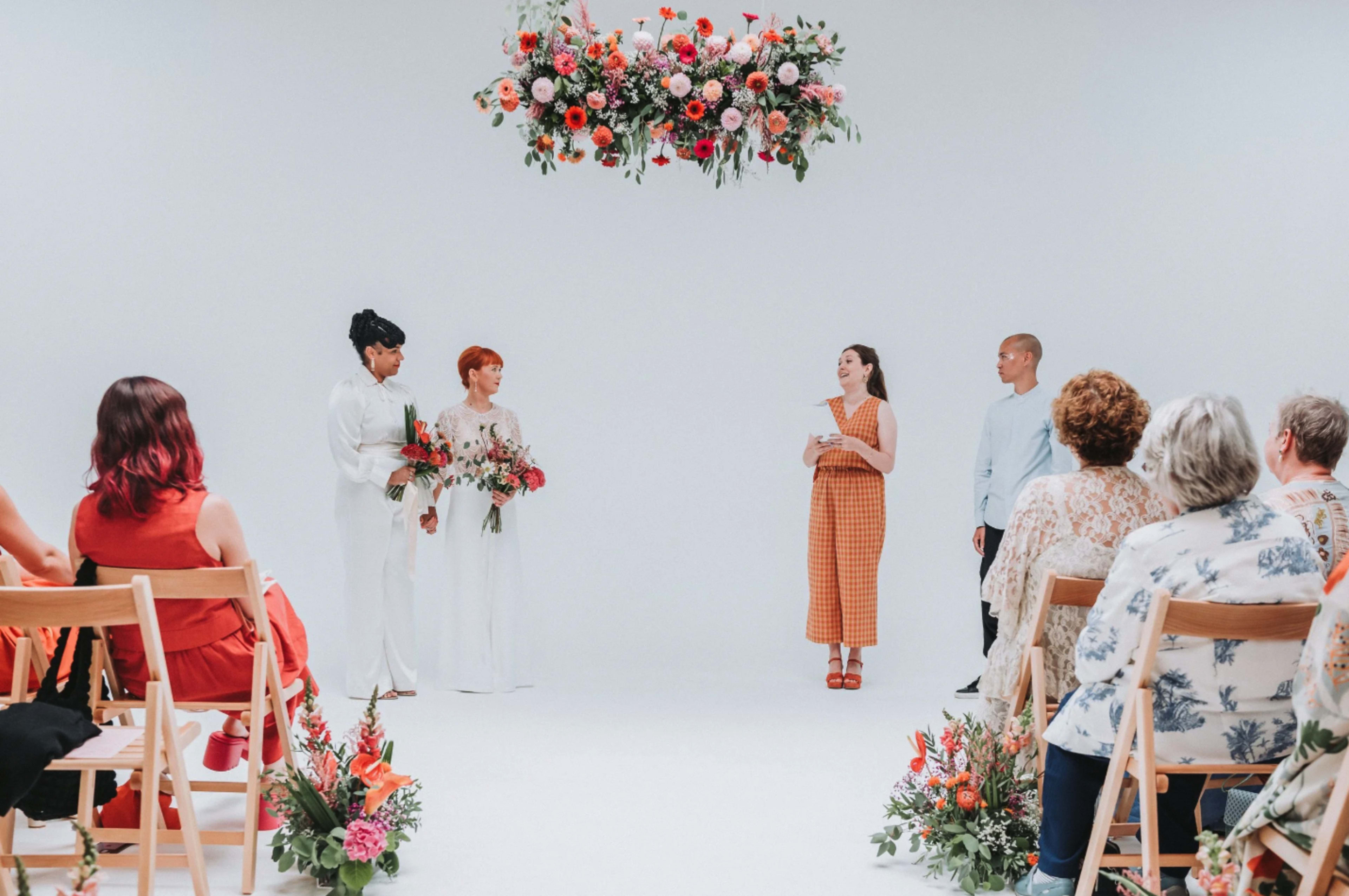 A wedding ceremony takes place in a minimalist white space, featuring a couple with floral arrangements, a speaker, and seated guests.