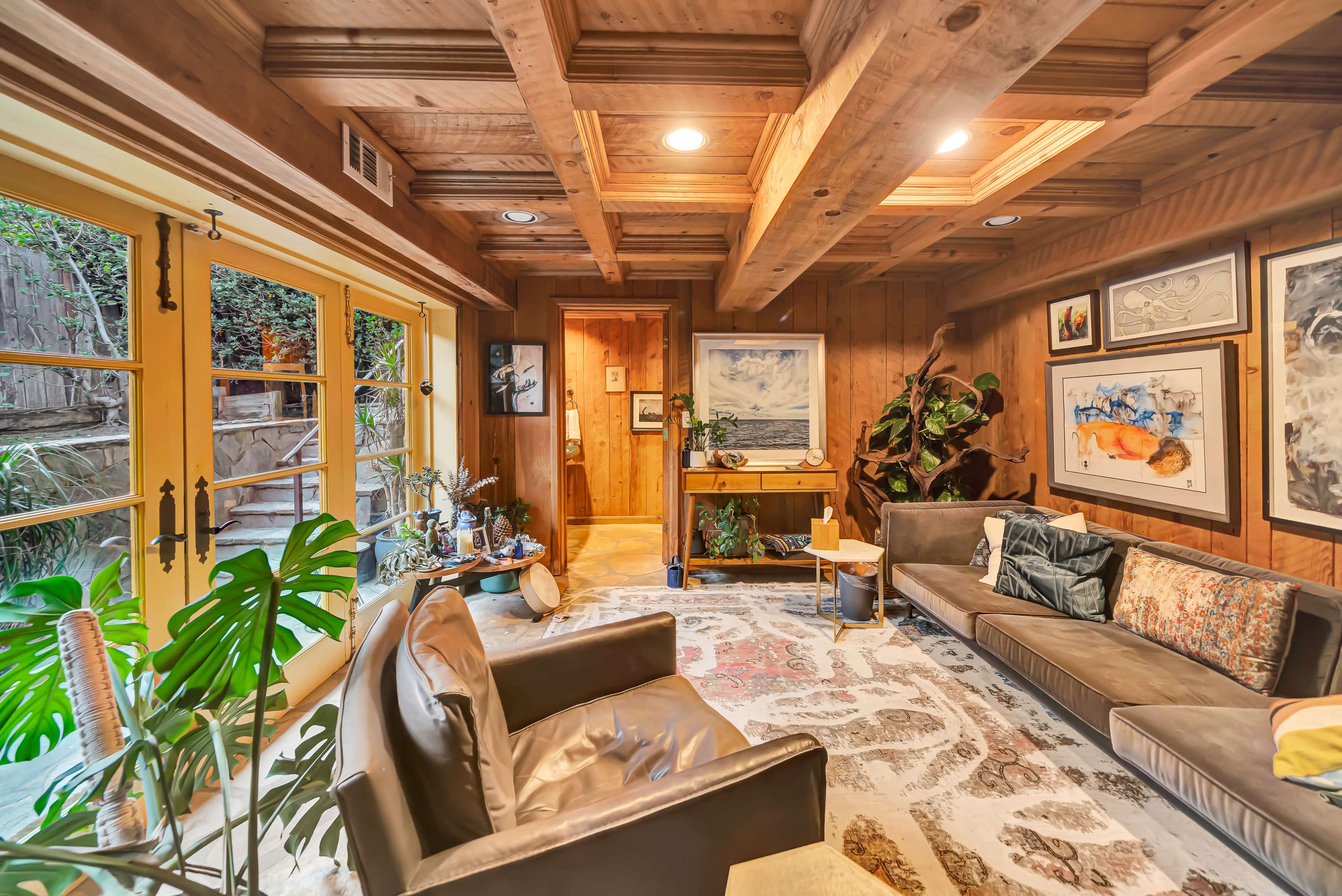 The image shows a cozy living room with wood-paneled walls, a large area rug, and framed artwork, featuring a sofa and a coffee table near large windows that overlook a garden.
