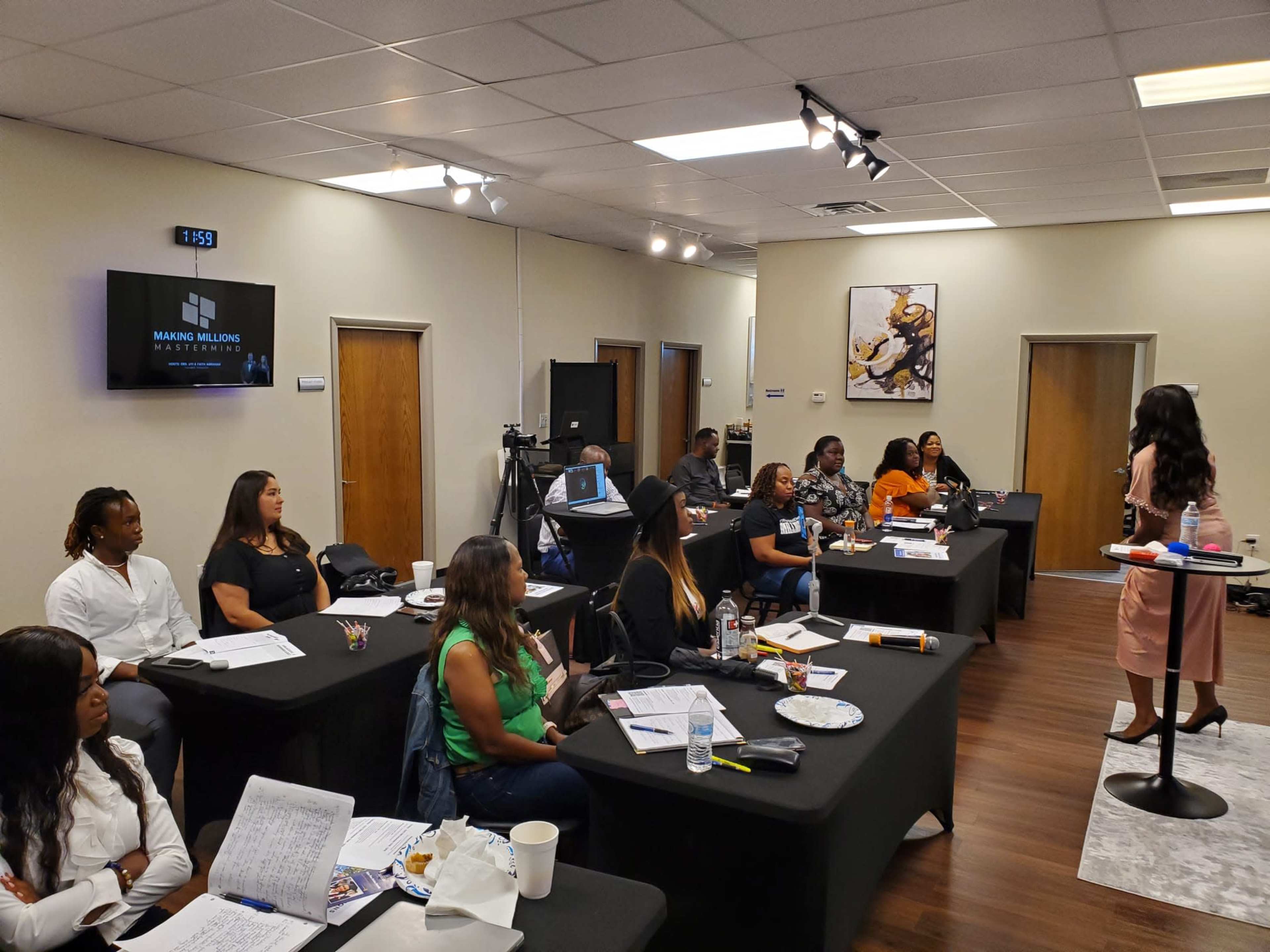 A diverse group of people attends a business presentation in a conference room, with a speaker addressing the audience while various materials are displayed on the tables.