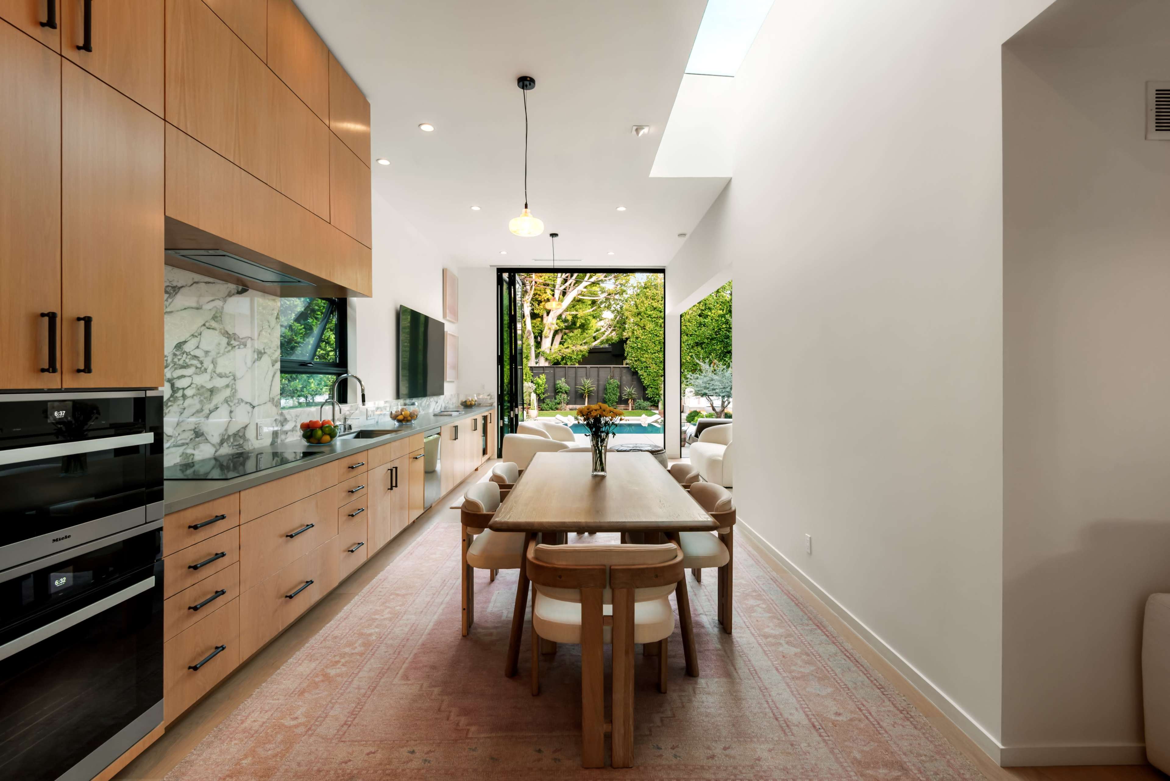 The image shows a modern kitchen with wooden cabinetry, a dining table, and large glass doors leading to an outdoor space.