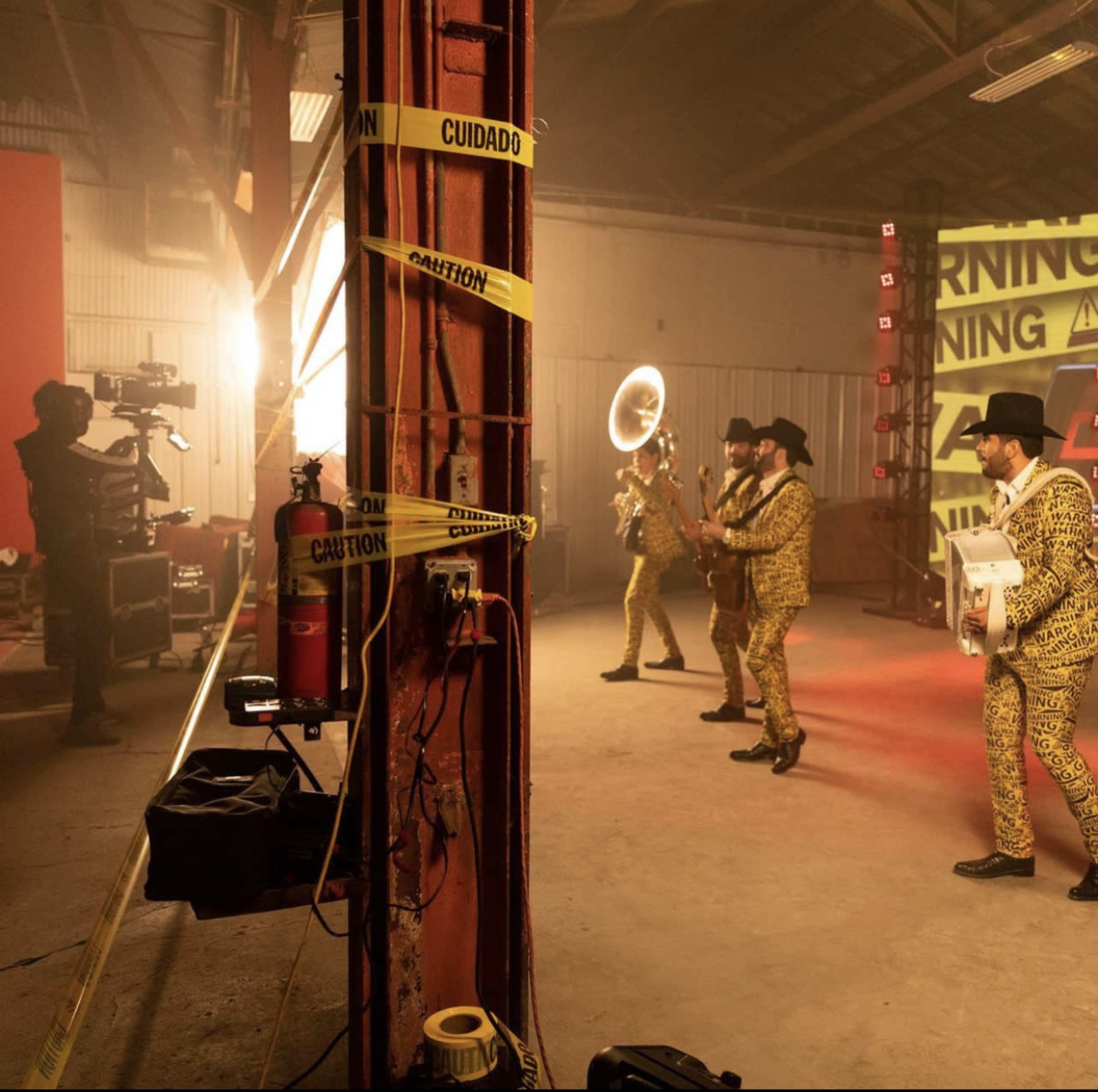A band in matching yellow outfits performs with instruments in a dimly lit warehouse decorated with caution tape and lighting equipment.