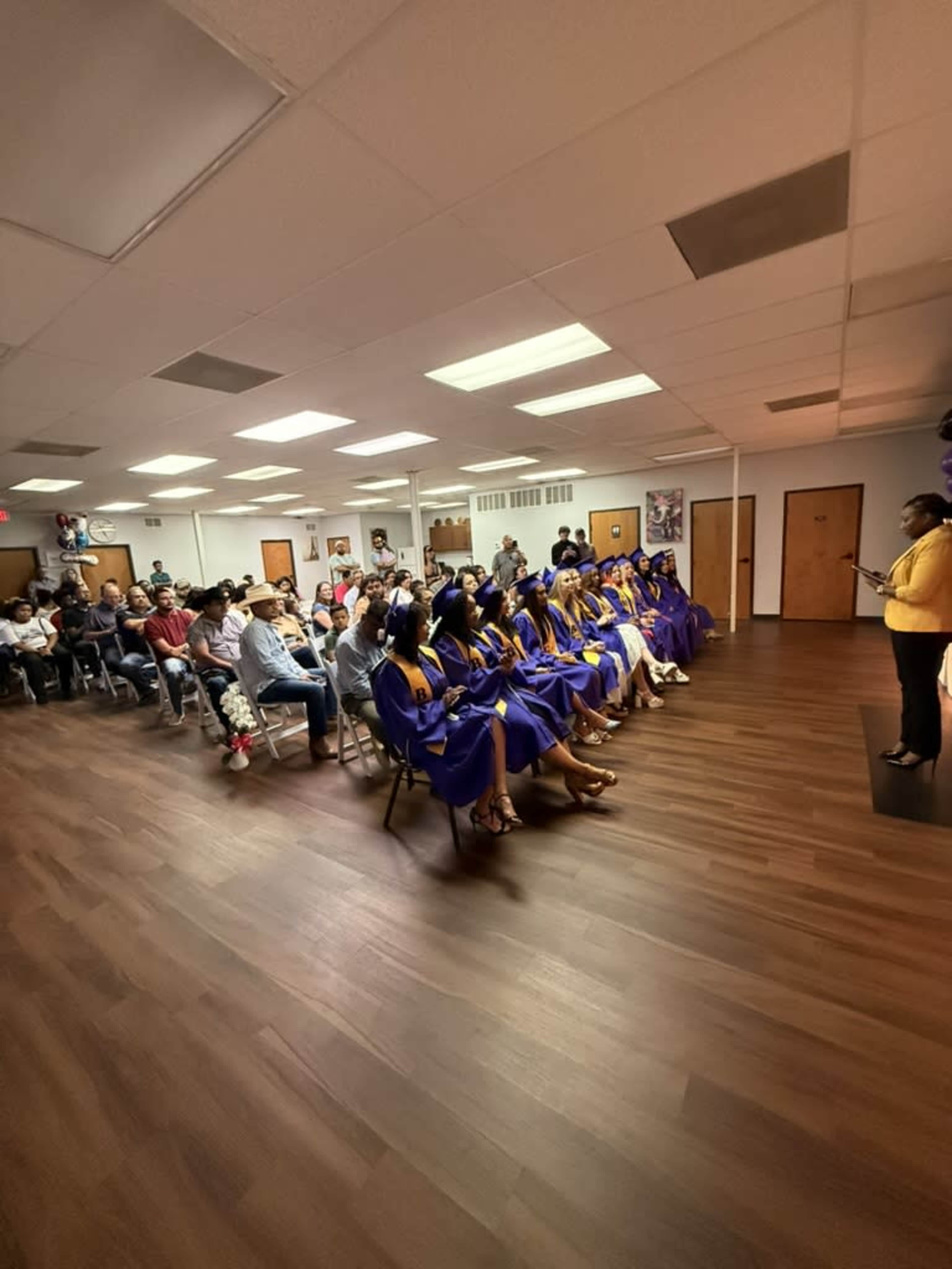 A group of graduates in blue gowns sits in rows facing an instructor during a graduation ceremony in a brightly lit room.