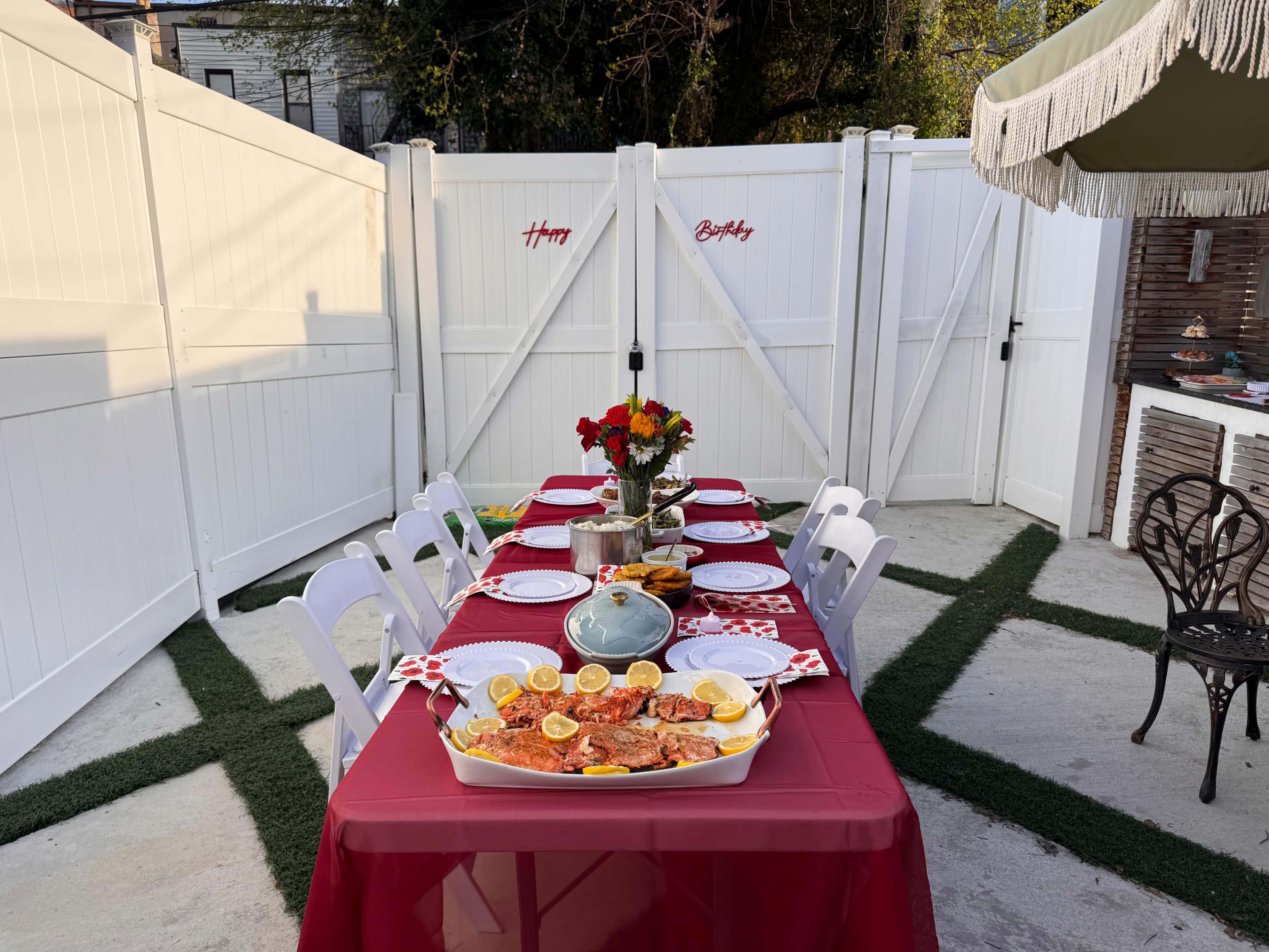 A long table is set with red tablecloth, plates, and a centerpiece of flowers, surrounded by a white fence and greenery in an outdoor space.