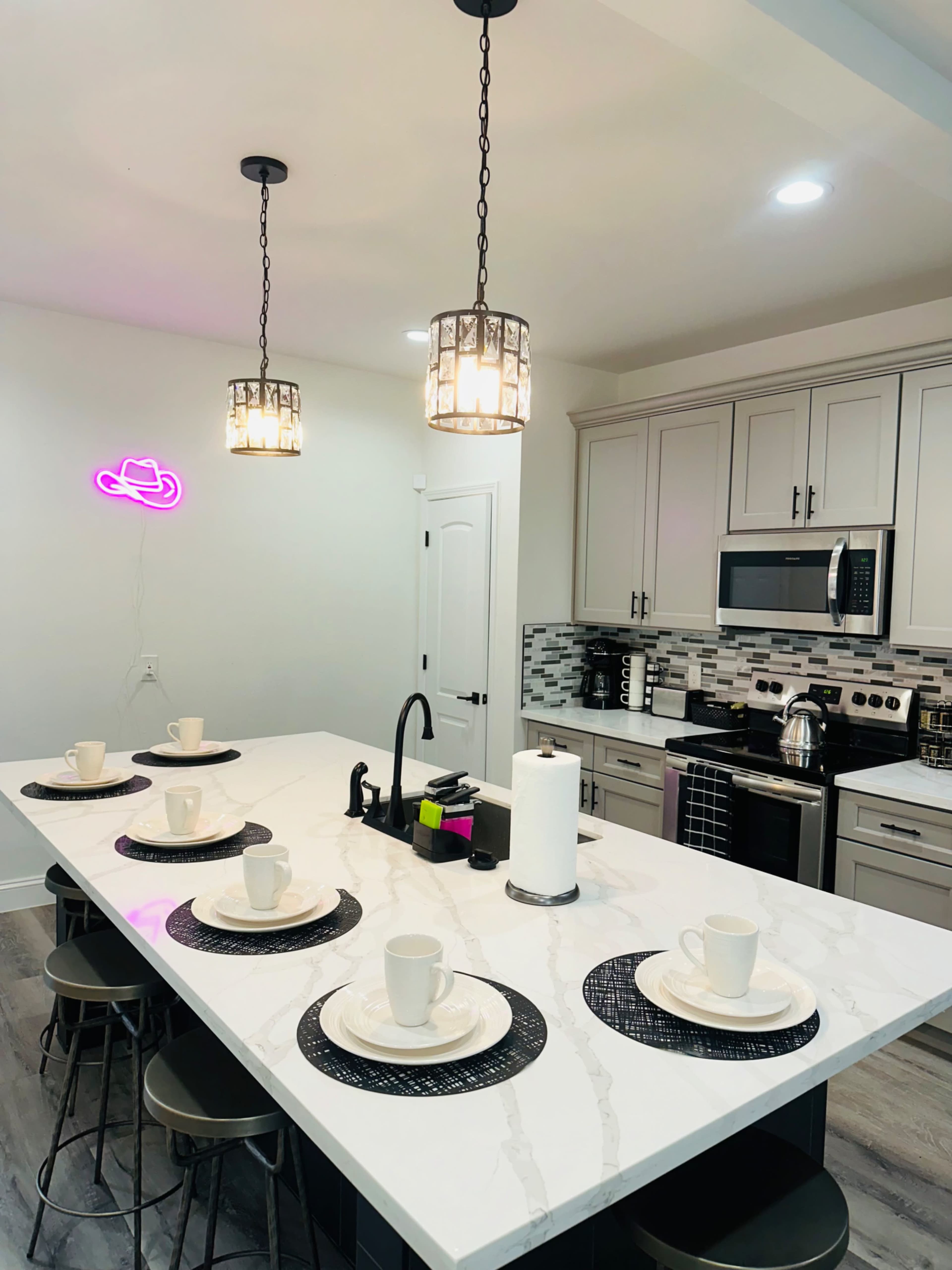 A modern kitchen features a long island with six plates and cups set on black placemats, illuminated by pendant lights and accented by a neon sign.