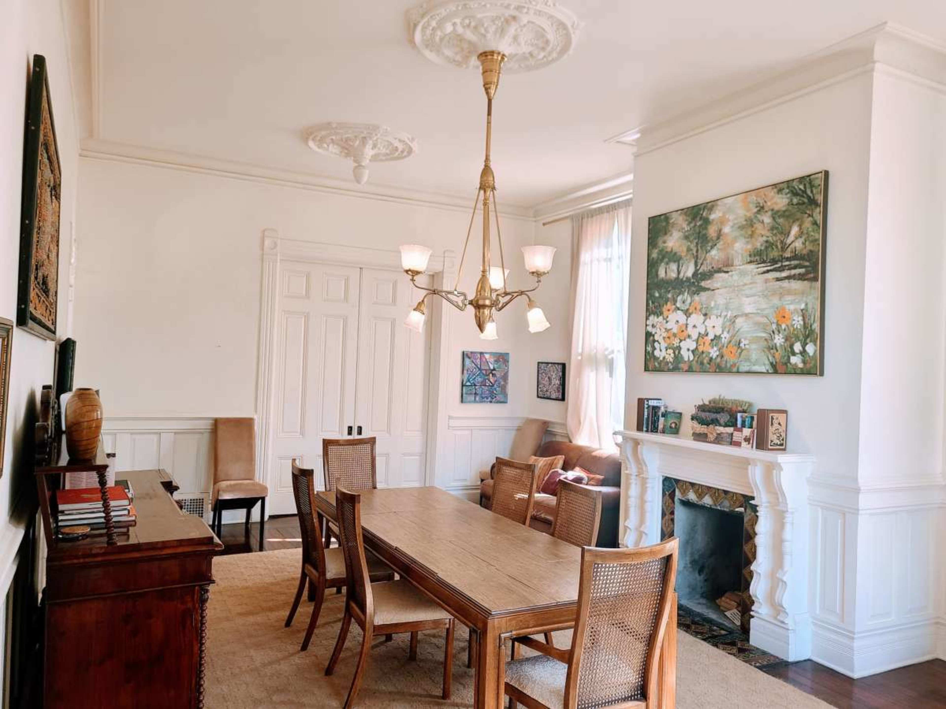 The image shows a bright dining area featuring a wooden table surrounded by chairs, a chandelier, and a fireplace with art pieces on the walls.
