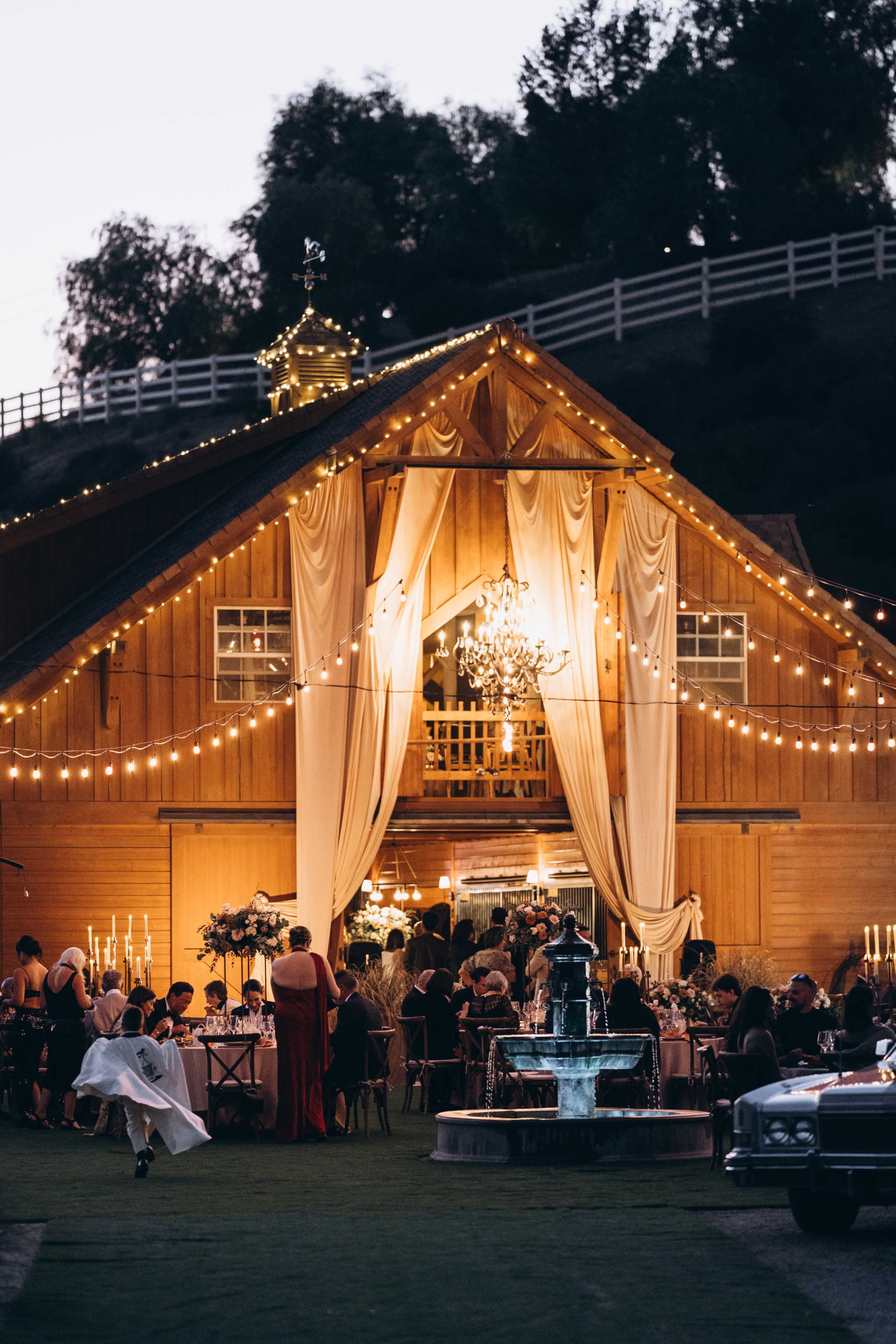 A beautifully decorated barn hosts an evening event with guests mingling under string lights and chandeliers, while a fountain is visible in the foreground.