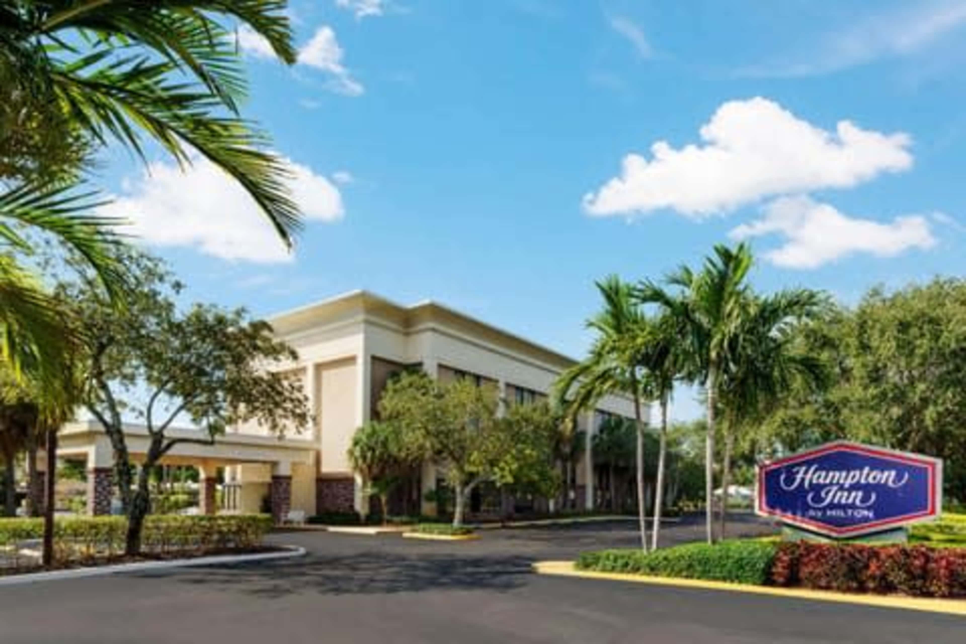 The image shows the exterior of a Hampton Inn hotel surrounded by palm trees under a clear blue sky.