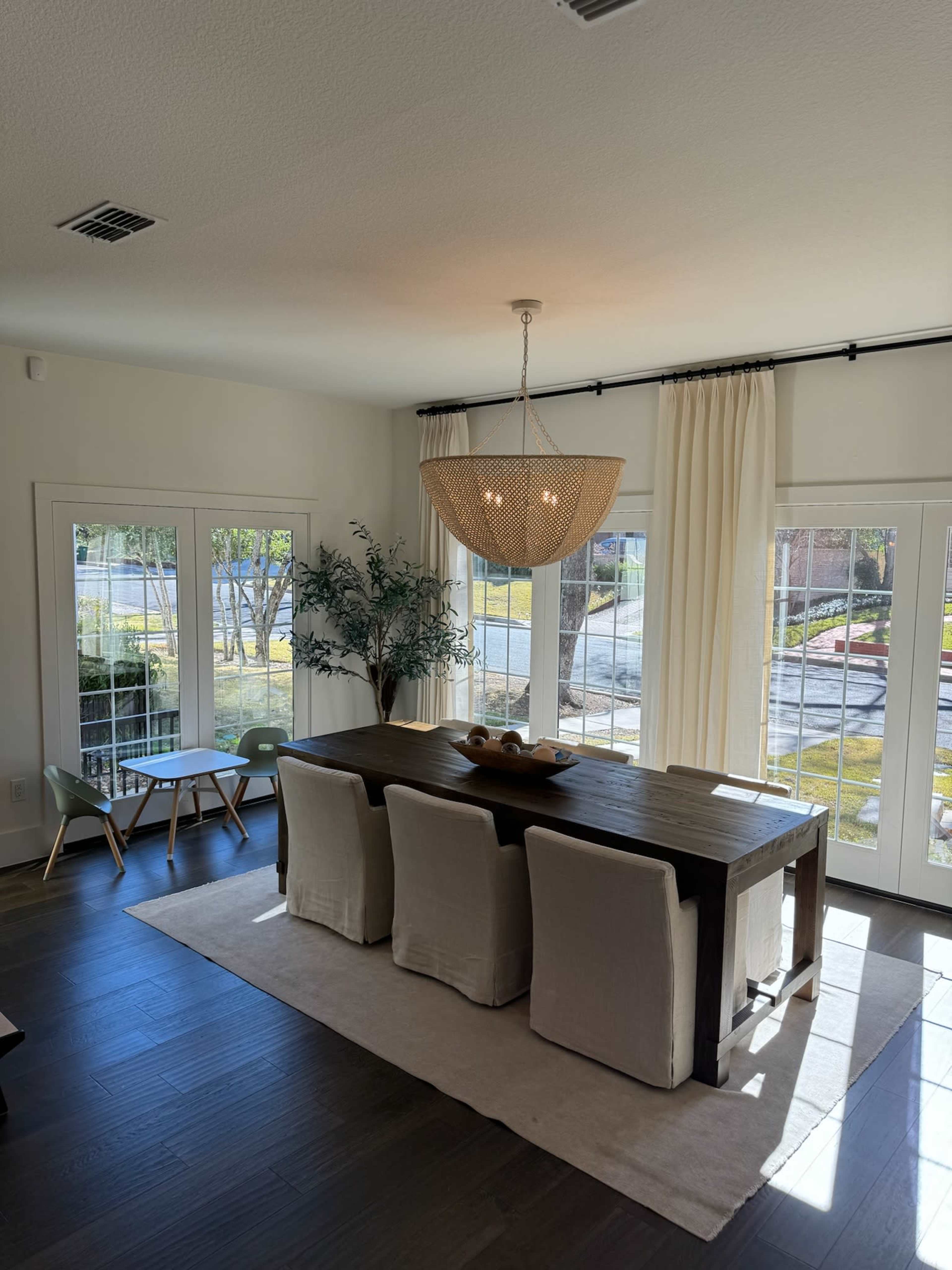 A dining room features a wooden table with six light-colored chairs, pendant lighting above, and large windows allowing natural light to fill the space.