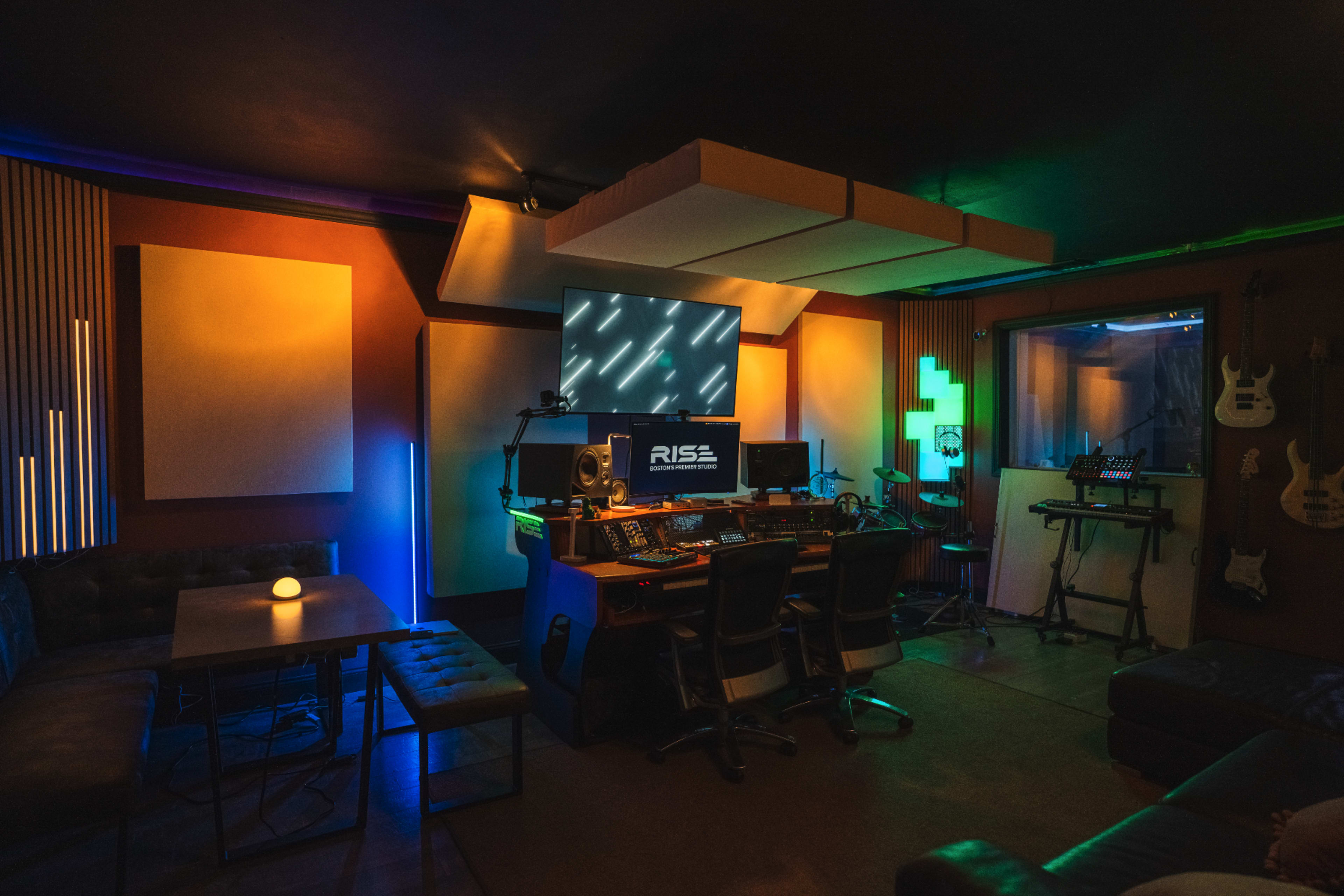 The image shows a modern recording studio equipped with a central mixing console, multiple instruments, and colorful ambient lighting.