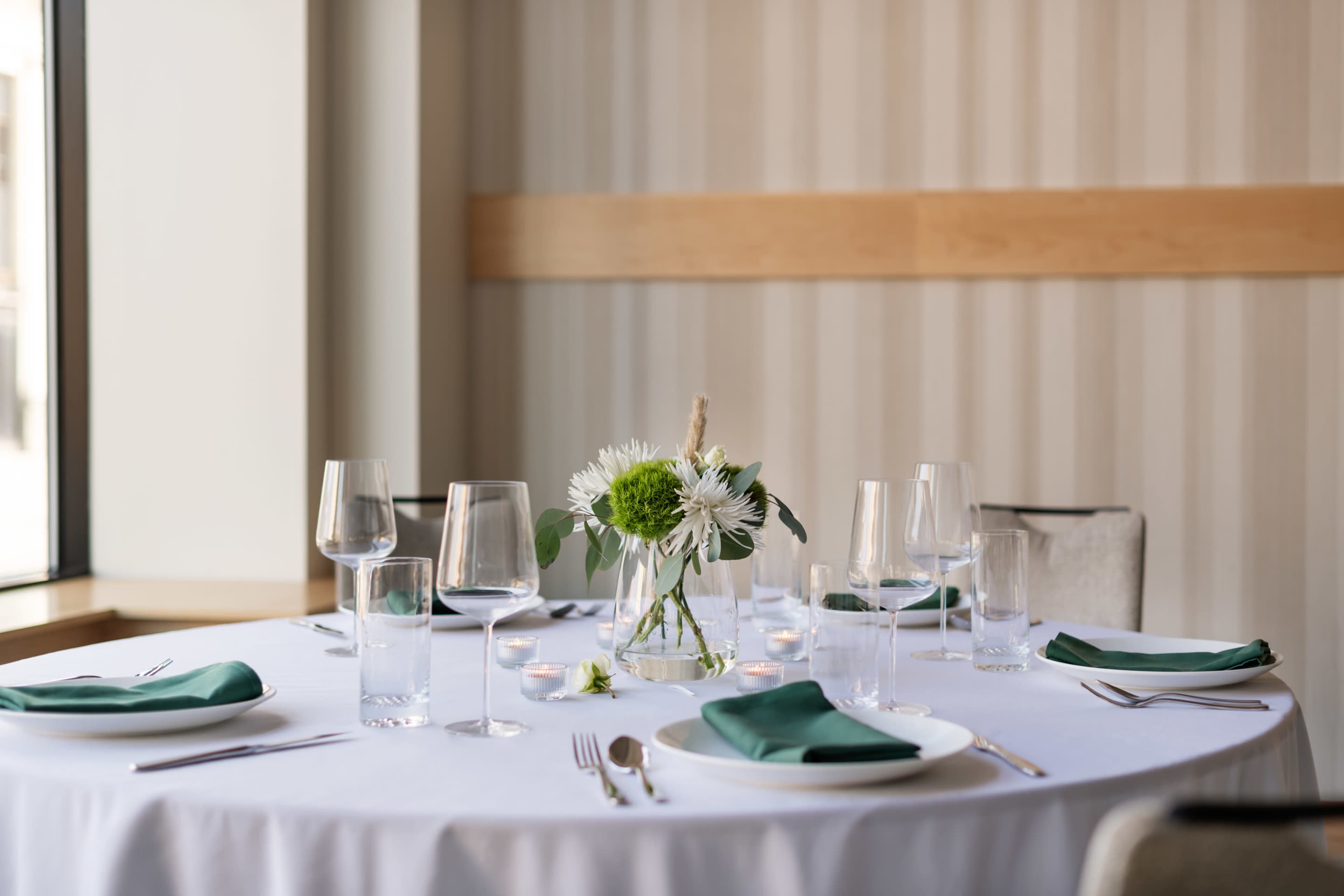 A round dining table is set with green napkins, glassware, and a floral centerpiece.