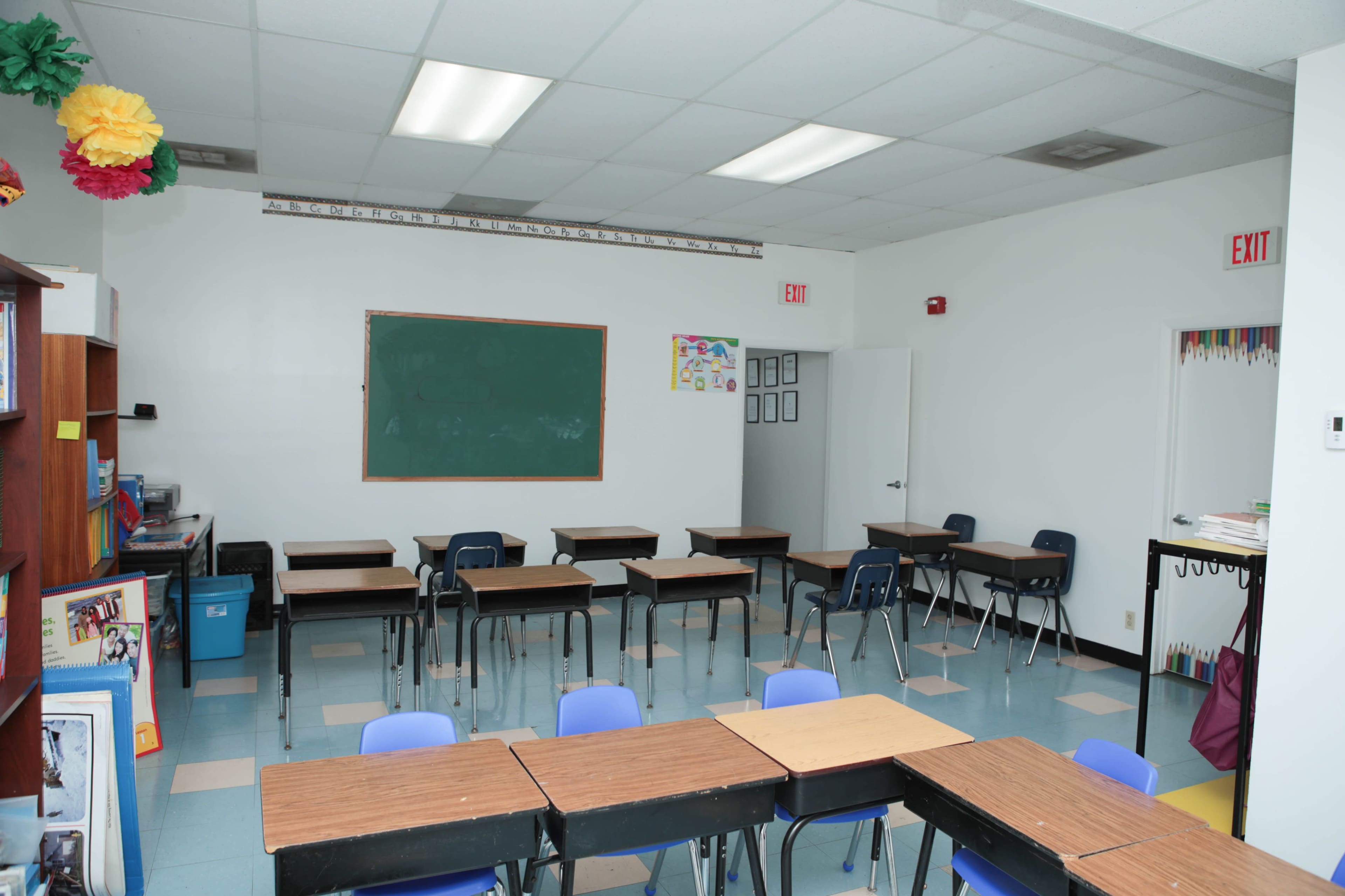 A classroom with several wooden desks arranged in a grid pattern, a green chalkboard on one wall, and a bookshelf on the side.