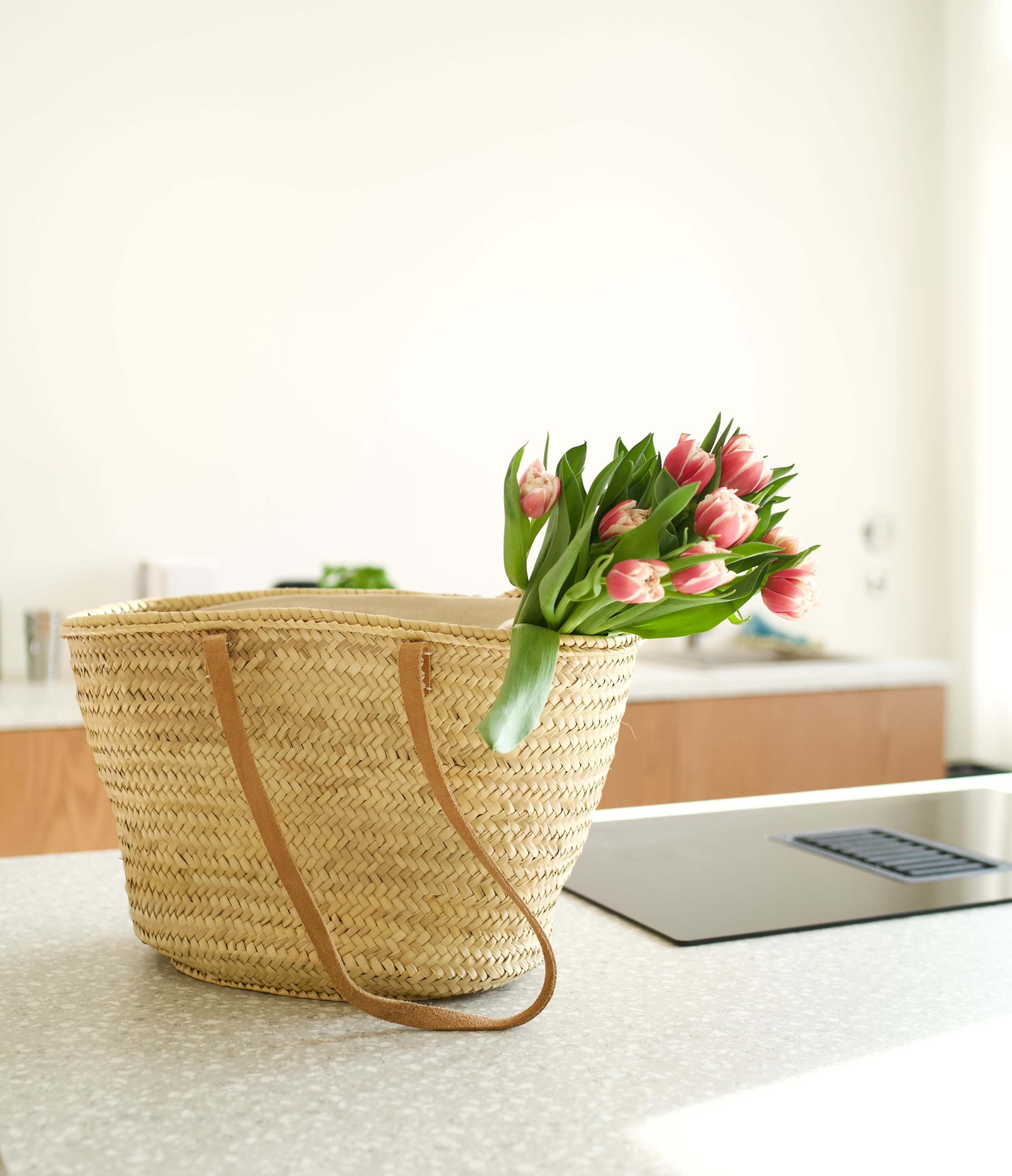 A woven straw bag with leather handles rests on a kitchen counter, holding a bouquet of pink tulips.
