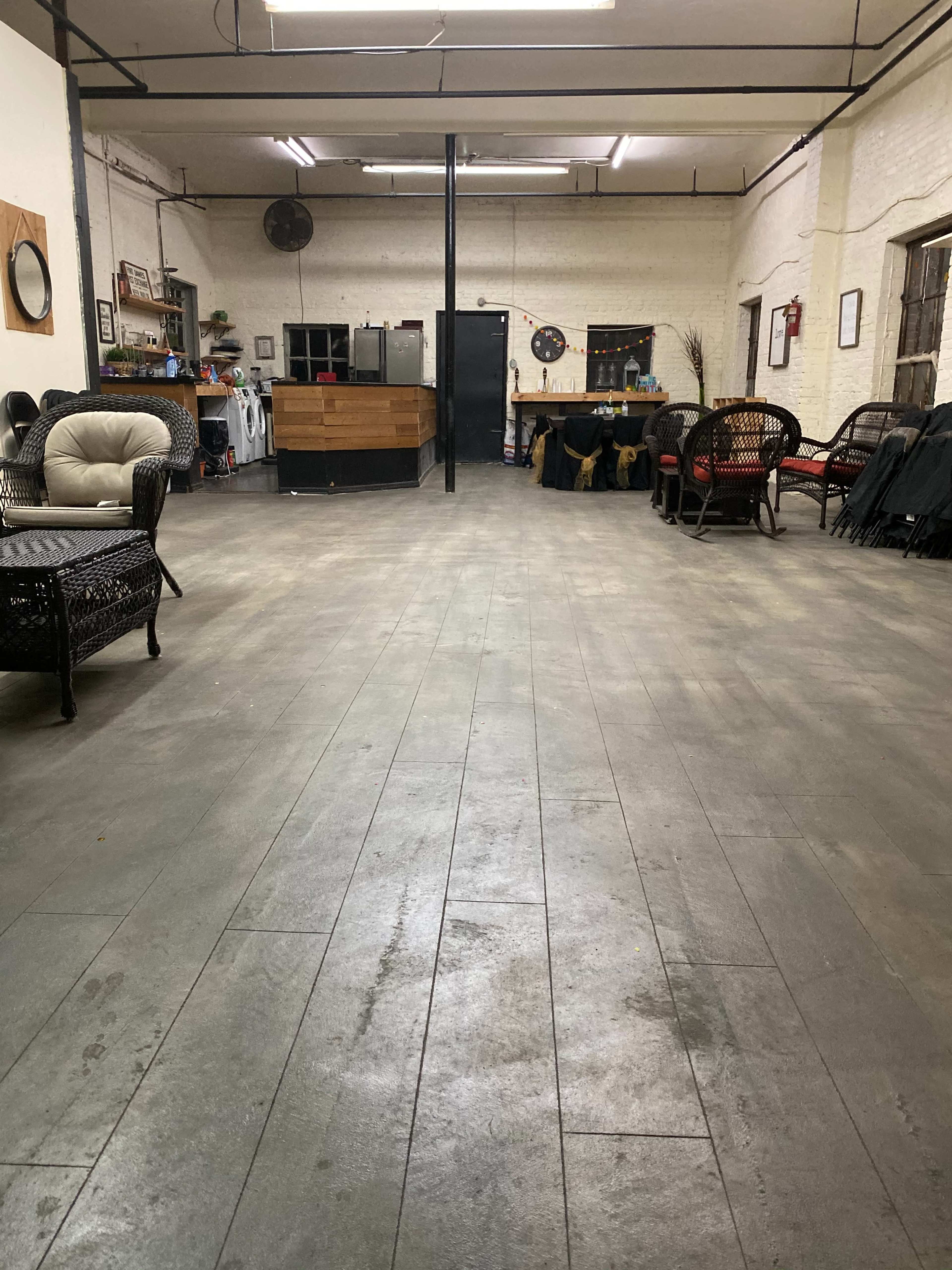The image depicts a spacious, empty room with wooden floors, featuring chairs on either side and a wooden counter in the background.