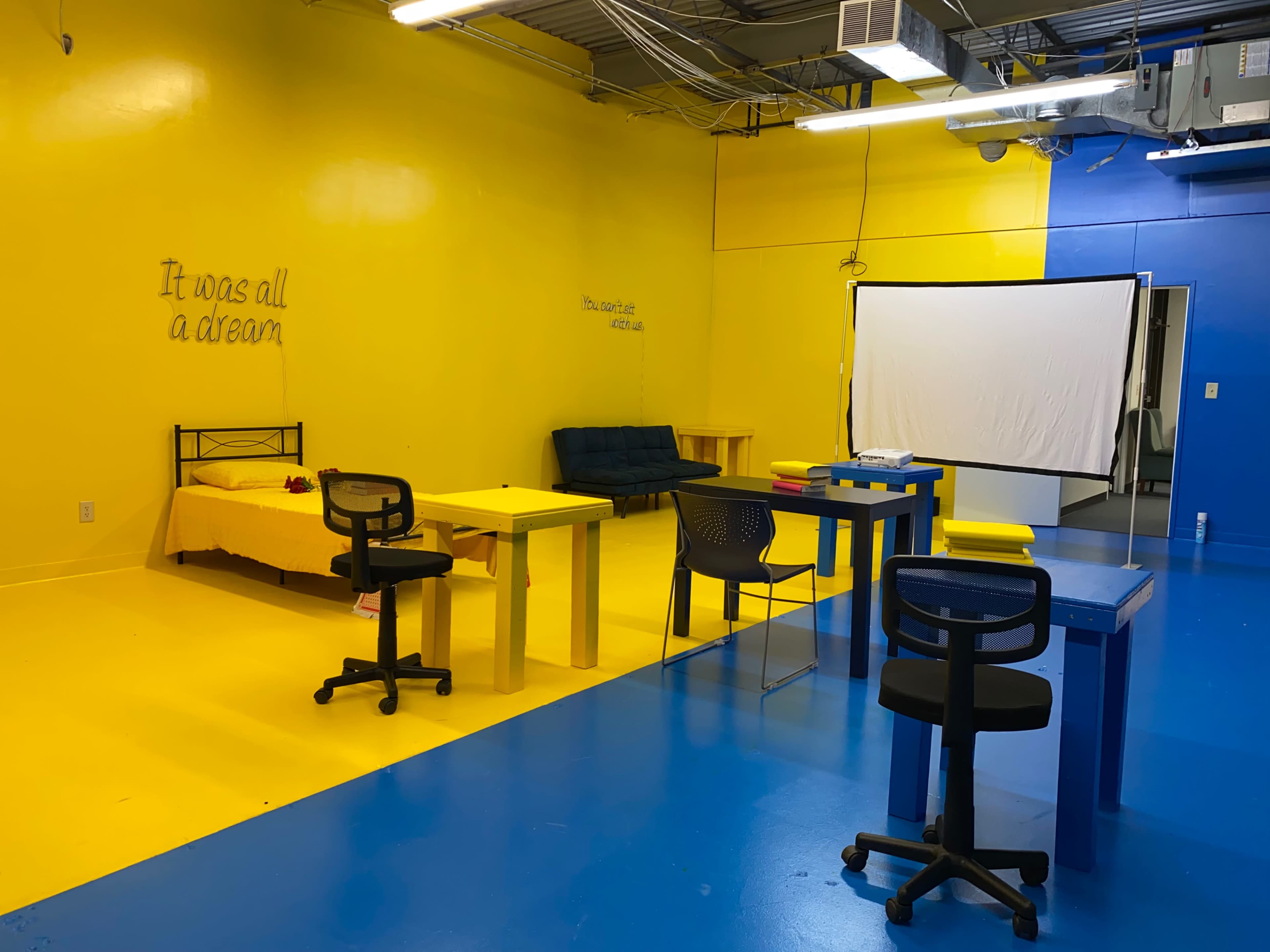 A brightly colored room with yellow walls and blue flooring, furnished with several tables and chairs, and a projection screen at one end.