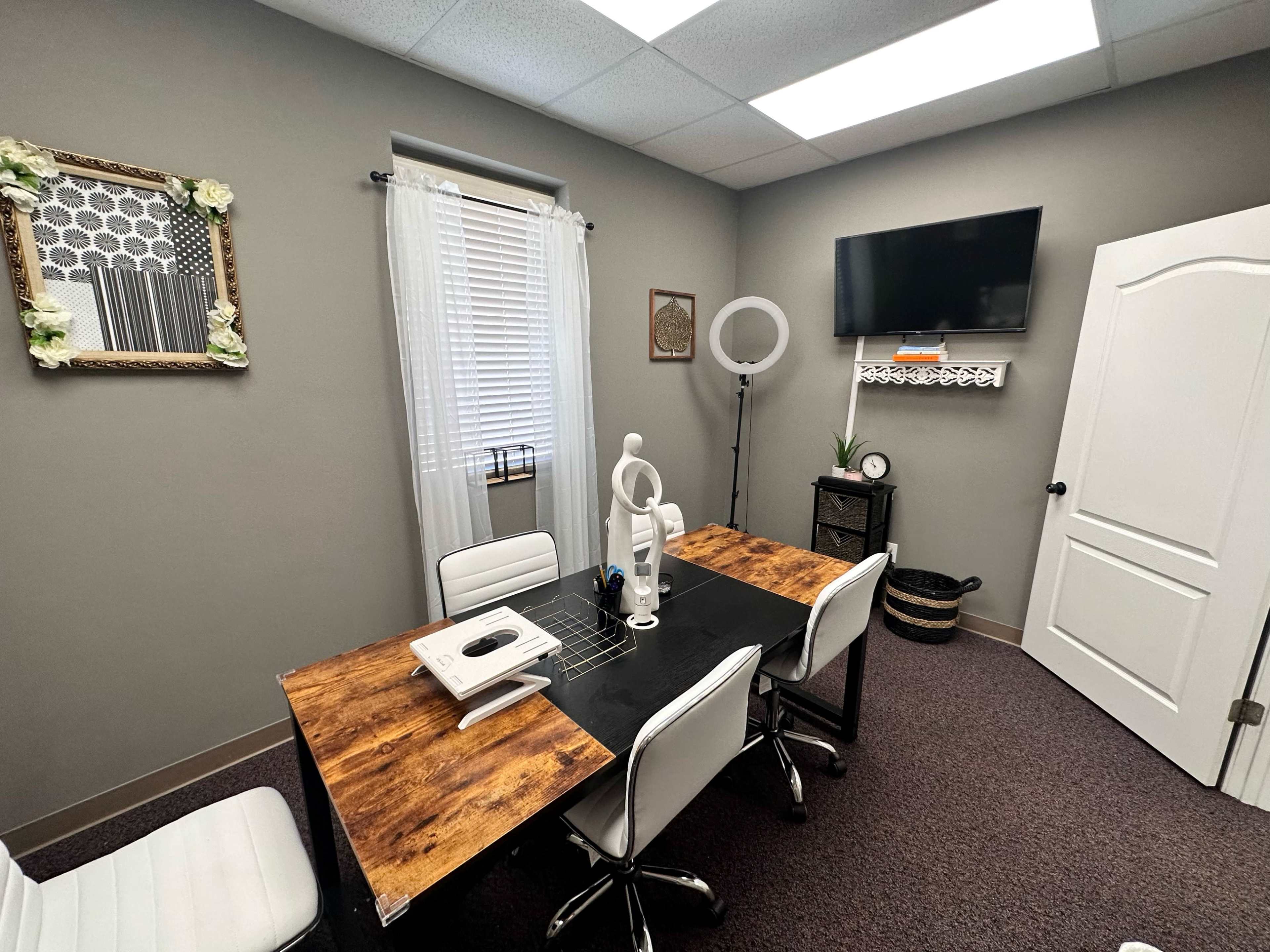 A small office space with a rectangular wooden table, four white chairs, a television mounted on the wall, and decorative elements like a framed artwork and plants.